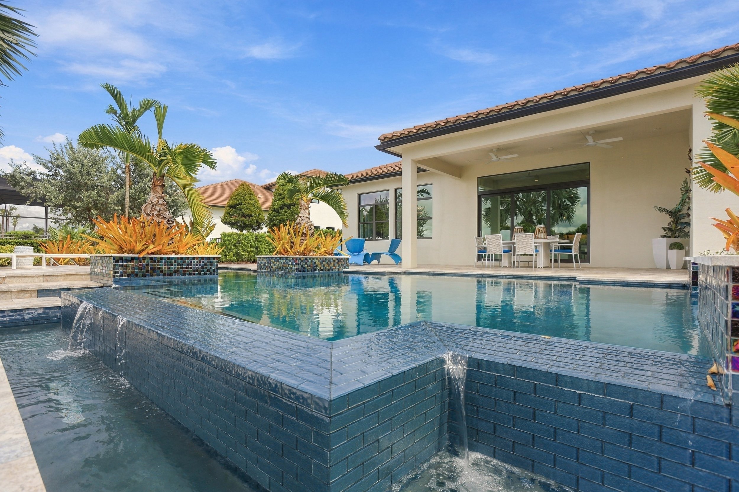 Blue tiled swimming pool with a small waterfall feature, surrounded by tropical plants and patio furniture under a clear blue sky.