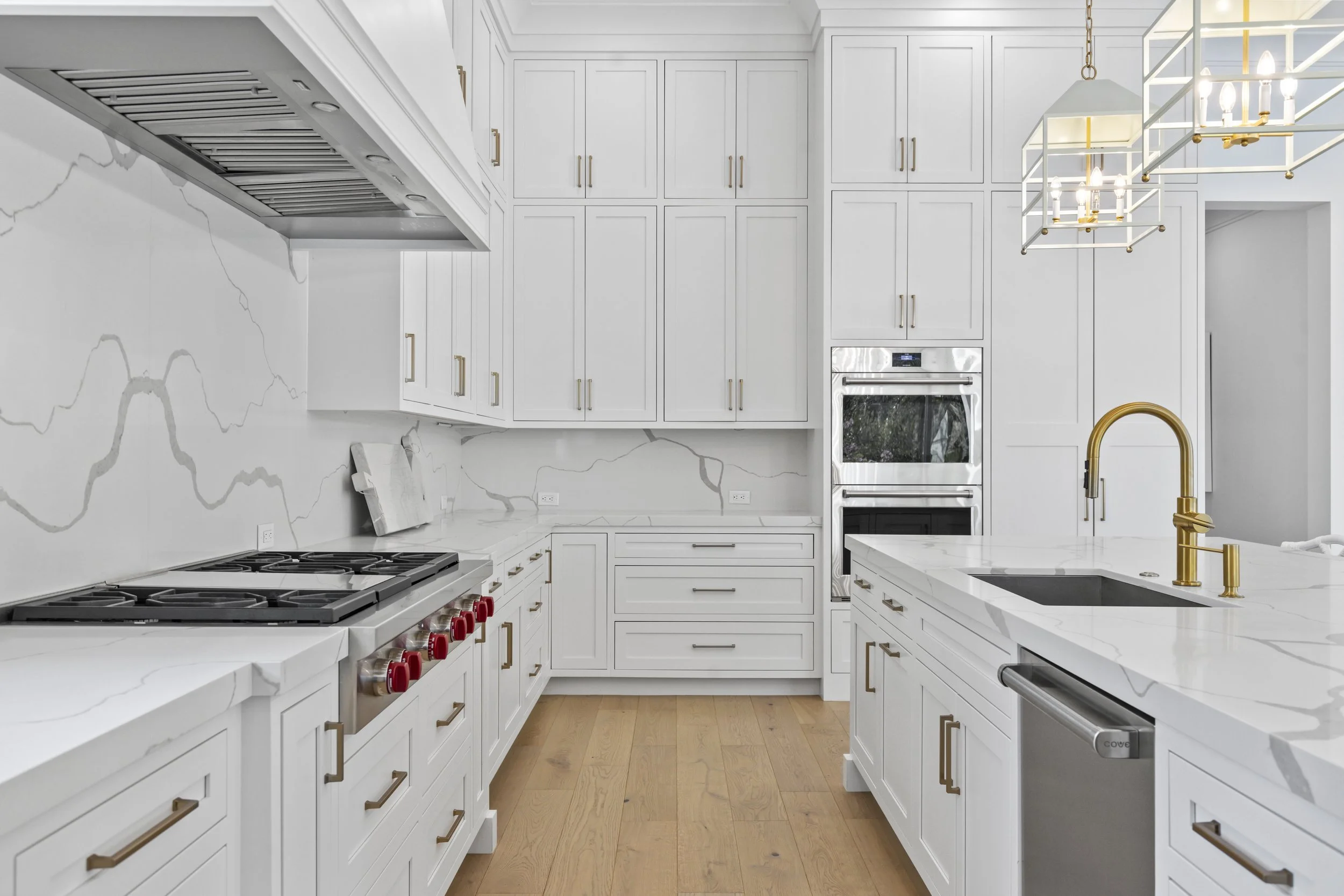 Modern white kitchen with gold hardware, marble countertops, and a hanging chandelier.