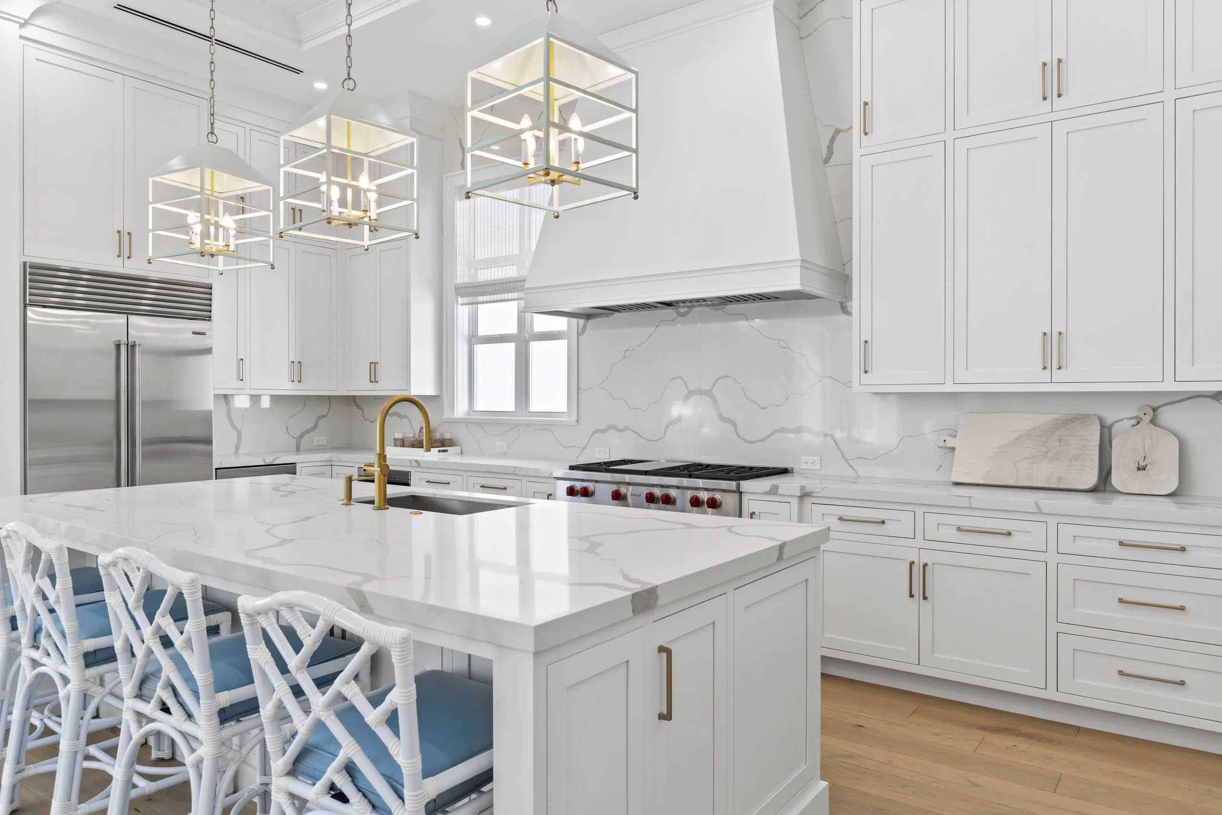A modern white kitchen with marble countertops, gold fixtures, and white cabinetry. Four square pendant lights hang above the island, which has a built-in sink. There is a stainless steel refrigerator on the left, and a window with white blinds above the stove.
