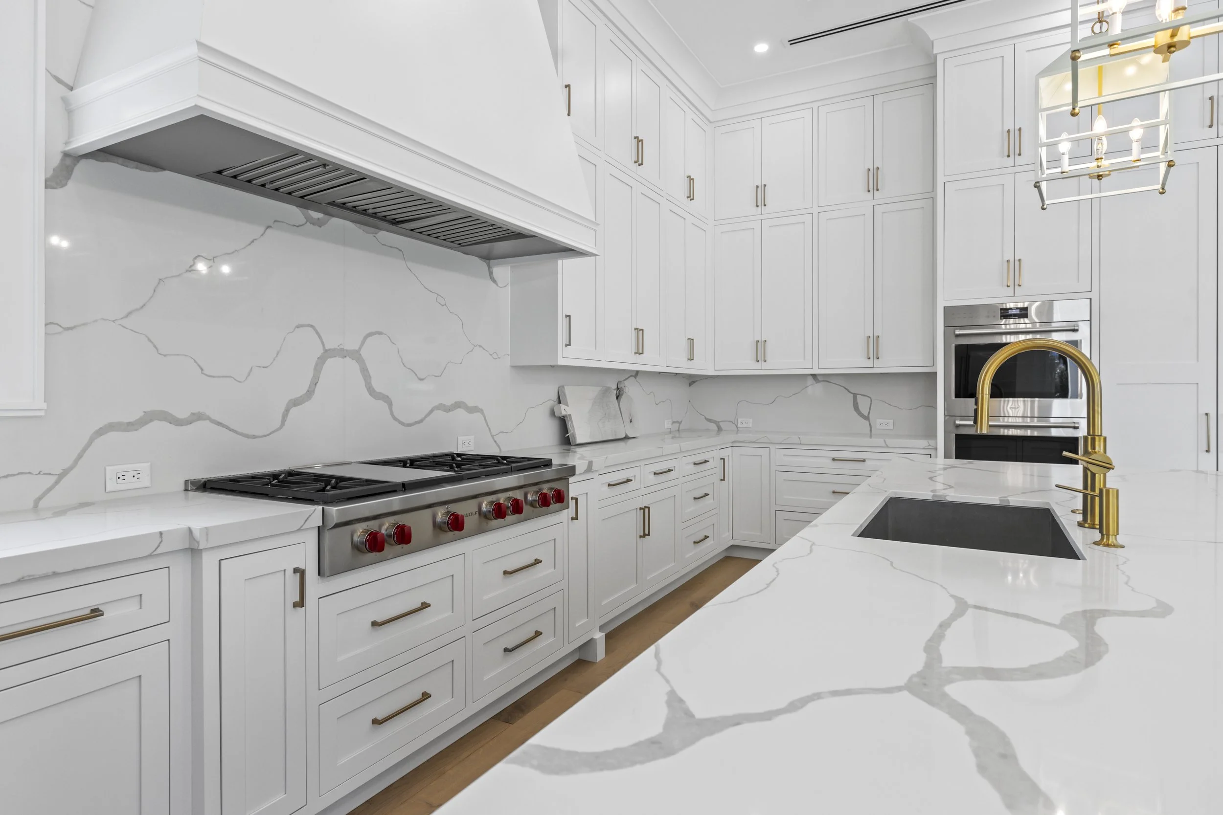 Modern white kitchen with marble countertops, gold hardware, and a stainless steel oven with red control knobs. There is a built-in microwave, a black sink with a gold faucet, and white cabinets with gold handles. A yellow chandelier hangs from the ceiling.