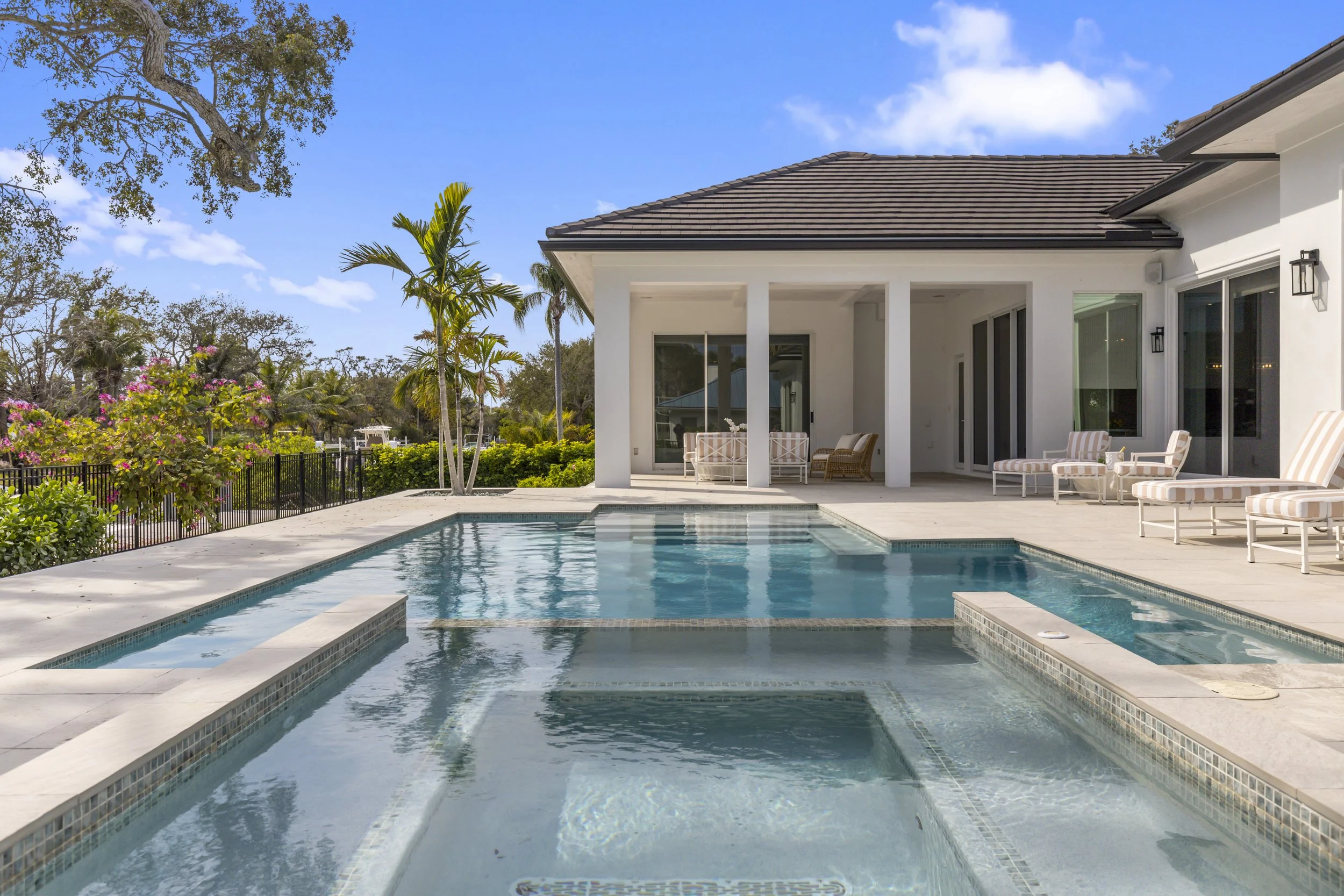 A backyard with a swimming pool, patio furniture, and a house with large glass doors, surrounded by trees and plants under a blue sky.