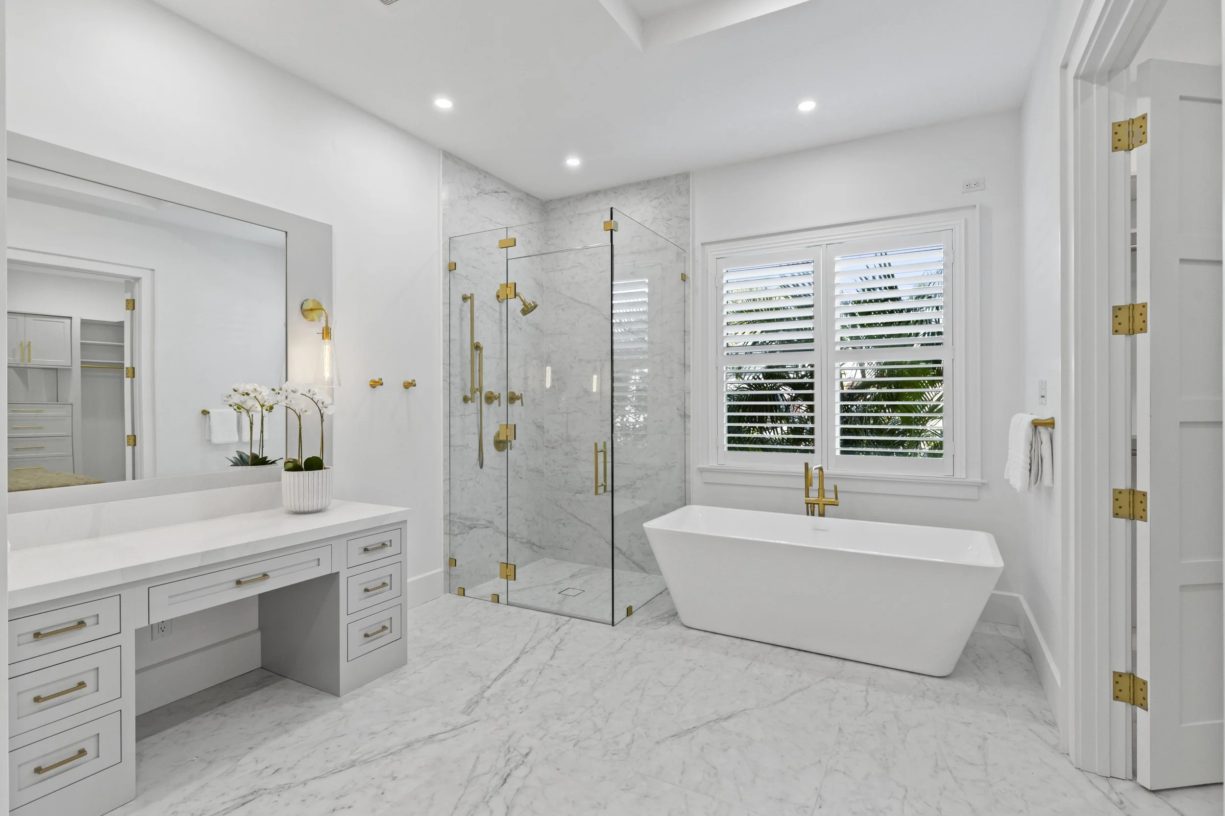 A modern, white bathroom featuring a freestanding bathtub, a glass-enclosed shower with gold fixtures, a white vanity with drawers and gold handles, and a window with white plantation shutters. There is also a white orchid in a pot on the vanity.