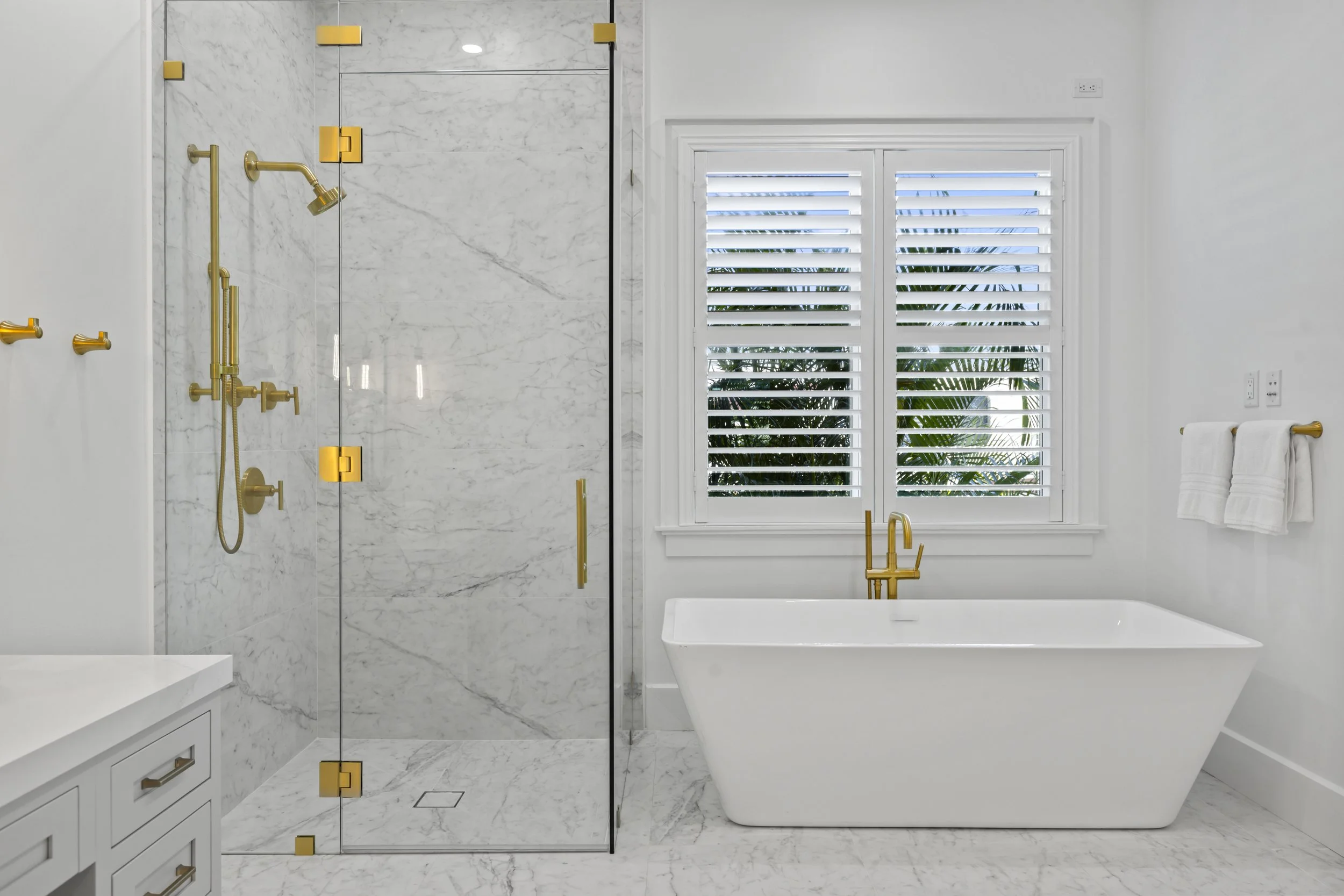 Modern bathroom featuring a glass-enclosed walk-in shower with gold fixtures, a white bathtub, a large window with plantation shutters, a white vanity with gold hardware, and white walls and marble flooring.