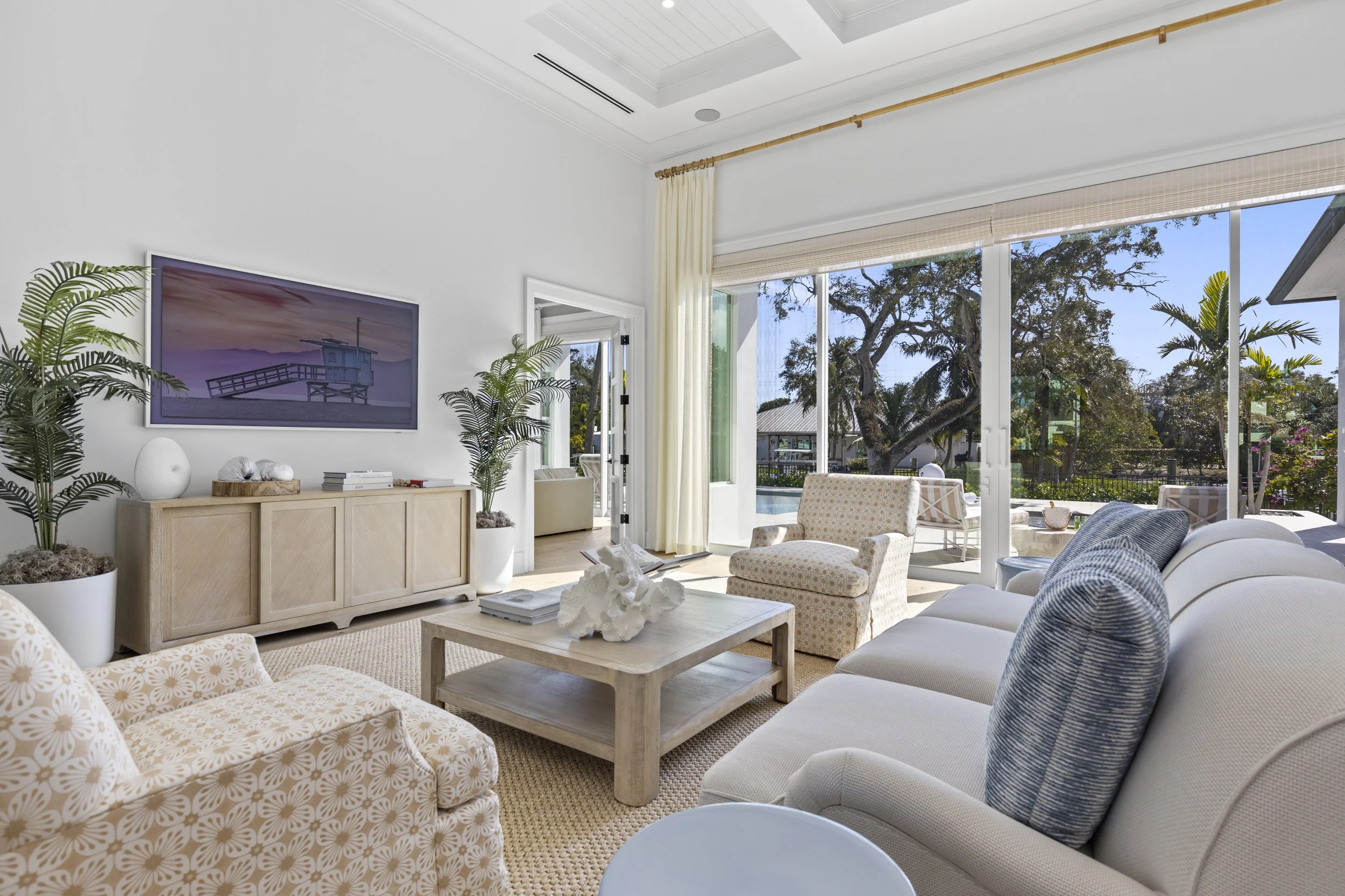 A bright living room with large glass doors opening to an outdoor area with trees and a pool. The room has white walls, beige curtains, and a variety of seating including a white sofa, patterned armchairs, and a wooden coffee table. Decor includes plants, a wall art of a lifeguard stand, and decorative objects.