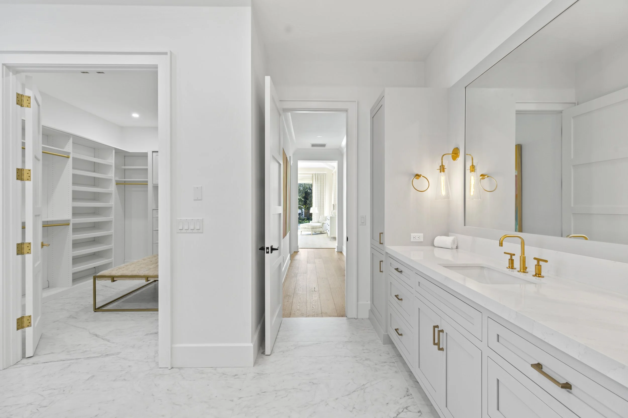 Modern bathroom with white cabinetry and gold fixtures, marble floors, and a view into a walk-in closet with white shelving and gold accents.
