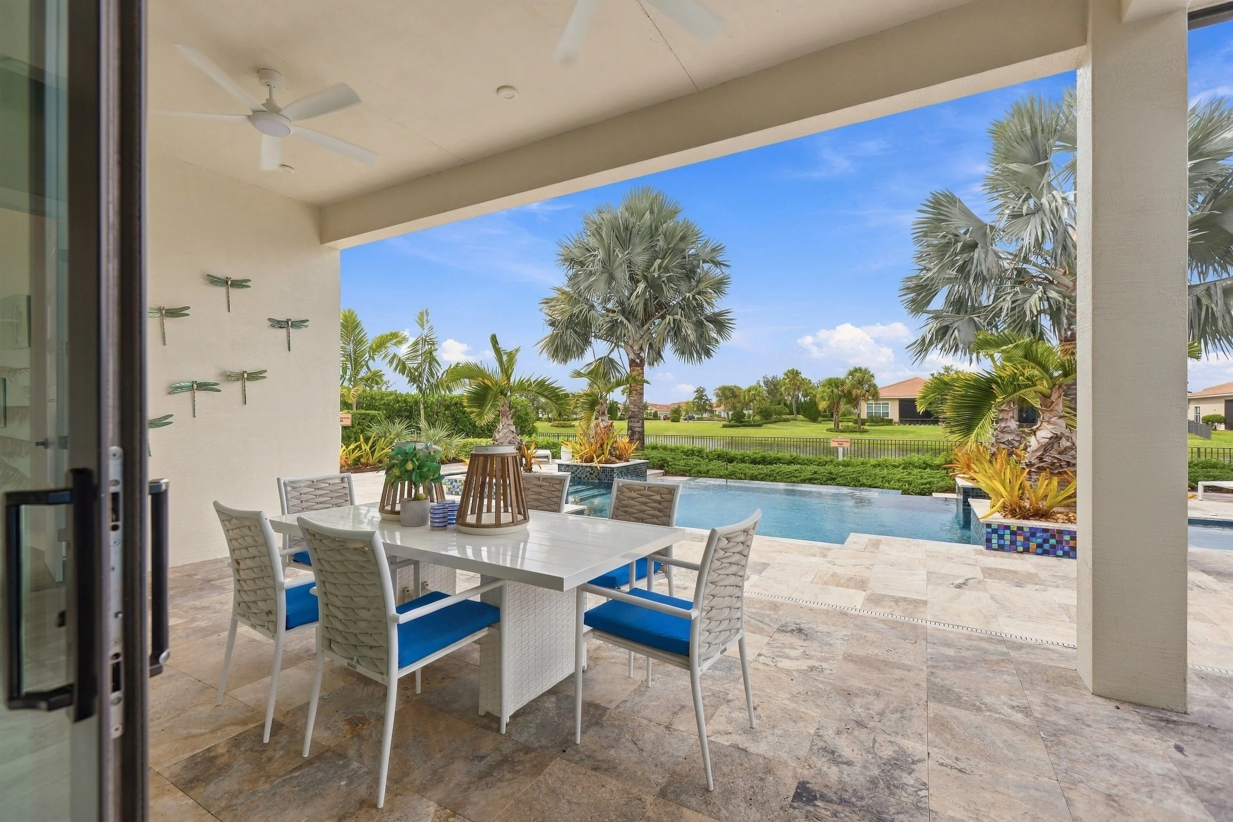 Image of a backyard patio with a white dining table and chairs, a swimming pool, palm trees, and a grassy yard with houses in the background, viewed from an open sliding door under a covered patio area with ceiling fans and dragonfly wall decorations.