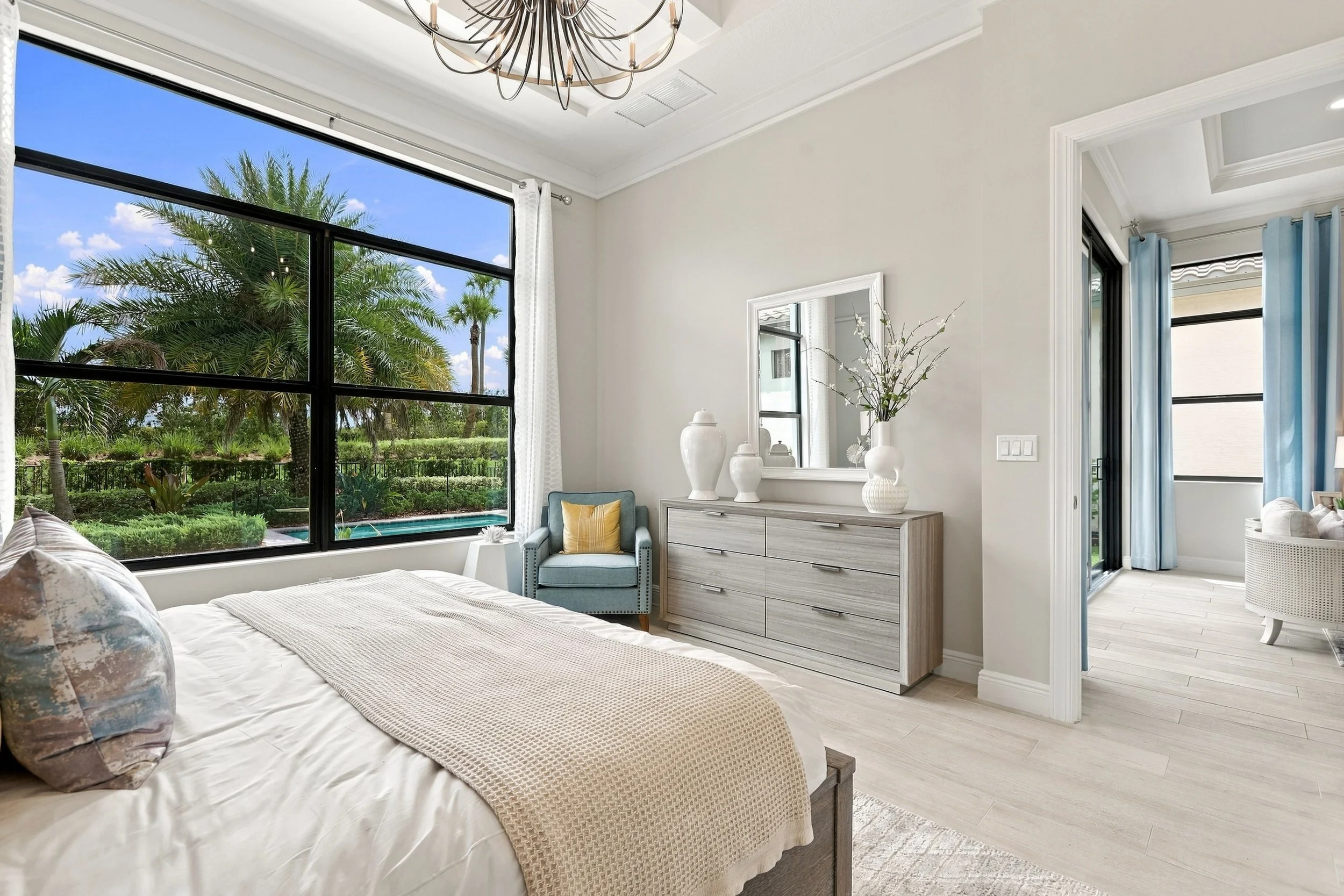 A bright bedroom featuring large windows with outdoor palm trees, a bed with decorative pillows, a blue armchair with a yellow pillow, a dresser with white vases and a mirror, and a sliding glass door with light blue curtains.