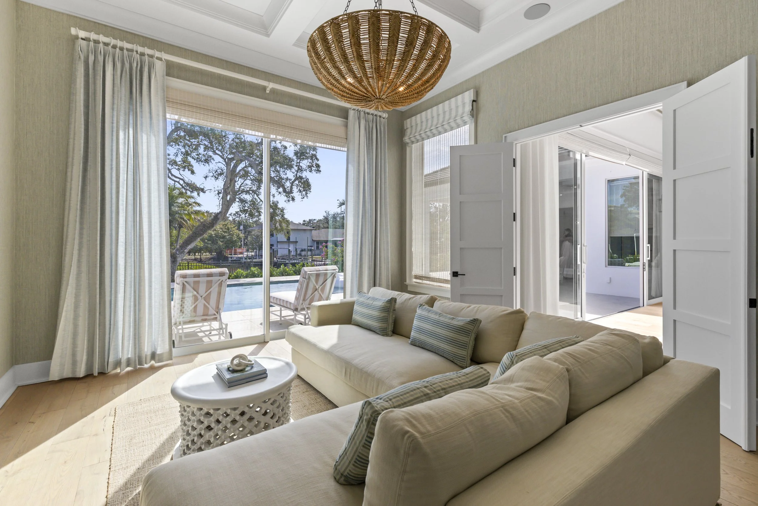 Living room with light-colored sofa and striped pillows, round white side table with decorative object and books, large window with sheer curtains and blinds, sliding glass door leading to patio with outdoor chairs, tree and buildings outside, floral chandelier hanging from ceiling.
