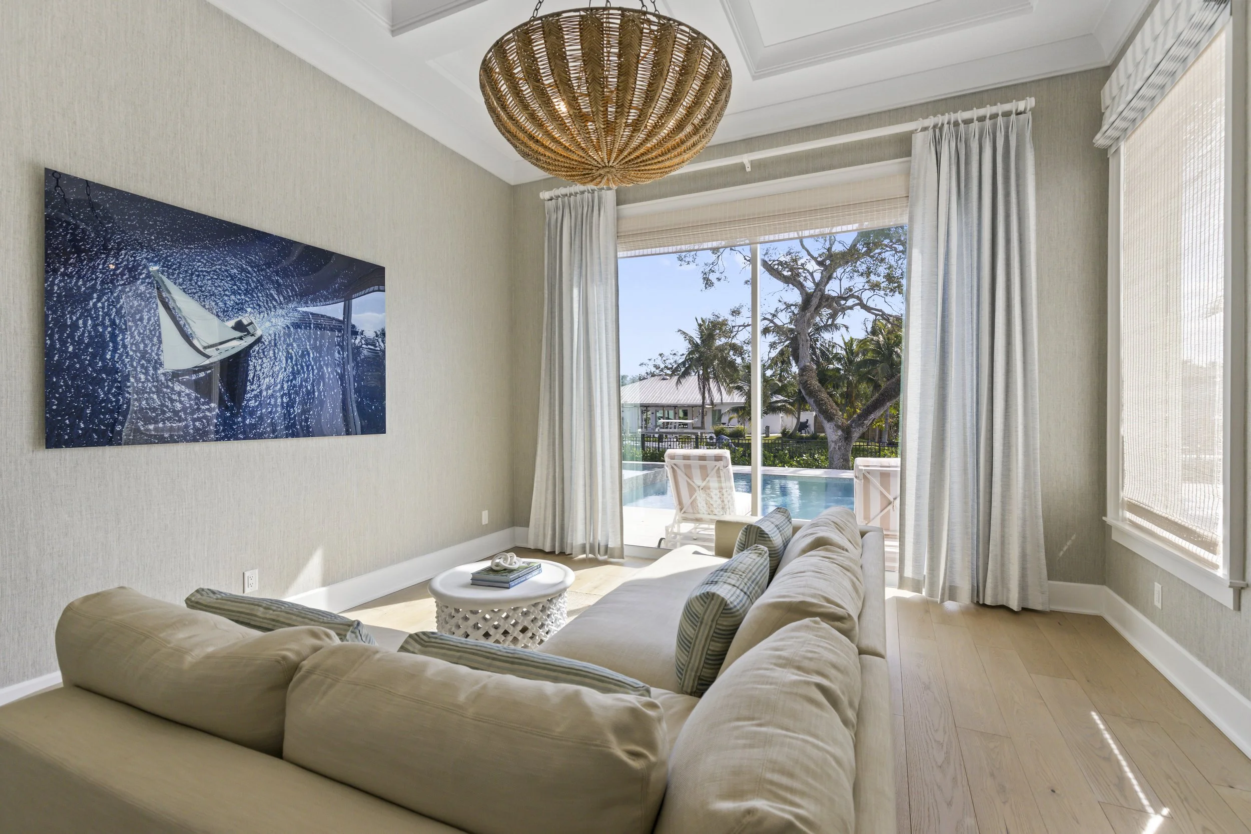 Living room with cream sectional sofa, striped pillows, round white coffee table with books, large window with white curtains showing trees and house outside, wall art depicting a sailboat, and a woven ceiling light.
