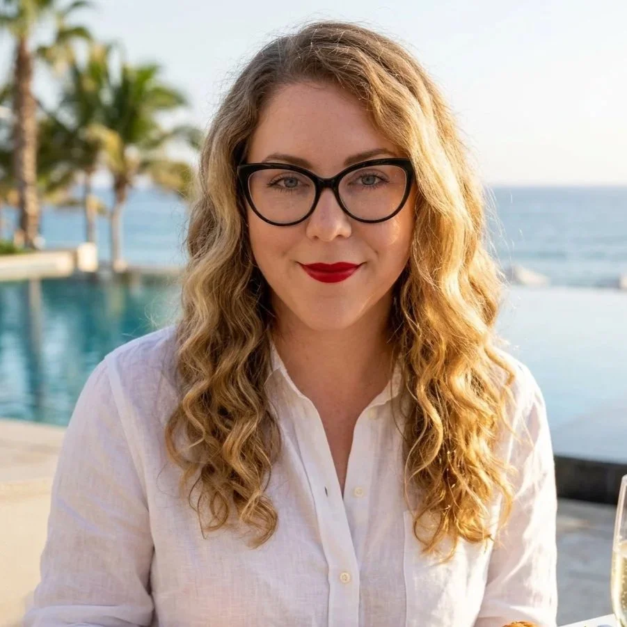 A woman with curly blonde hair, wearing glasses and a white shirt, smiling outdoors near a body of water with palm trees in the background.