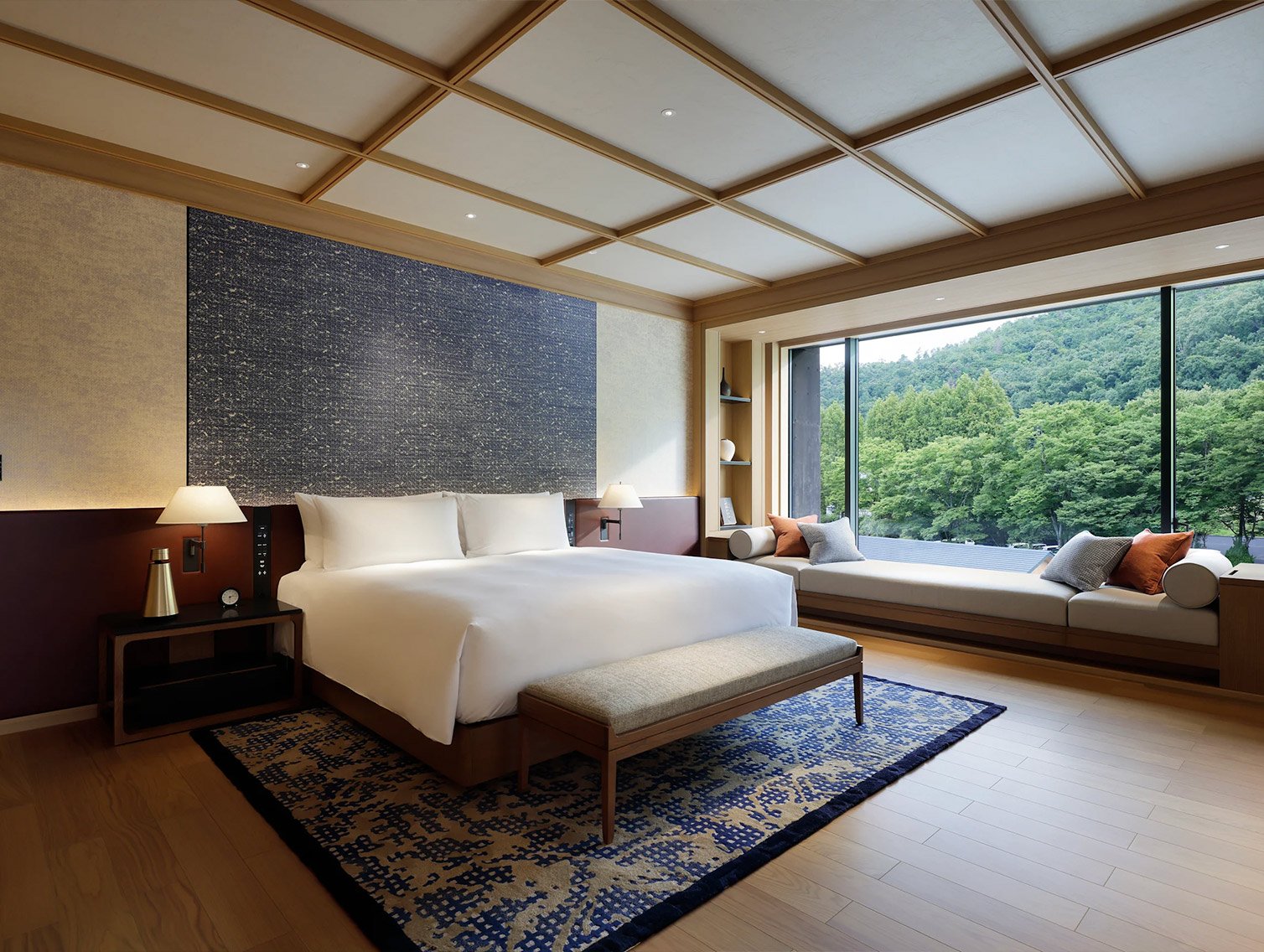 A modern bedroom with a large bed, bedside lamps, a window seat with pillows, a large window showing a green scenic view, and minimal decor.