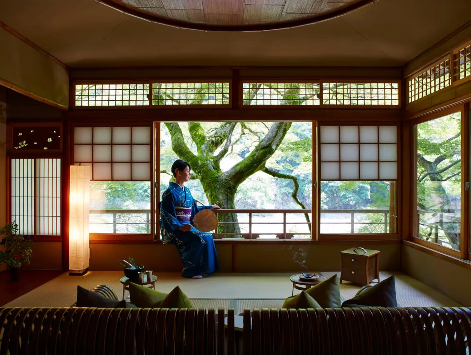 A woman in a blue kimono sitting on a window ledge in a traditional Japanese room with shoji screens, a paper lantern, and a small table with incense, overlooking a garden with a large tree.