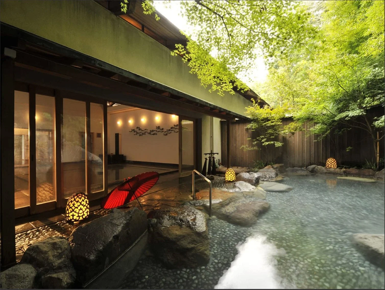 An outdoor Japanese hot spring bath (onsen) surrounded by rocks, lush green trees, and traditional Japanese lanterns, with a view of a building interior through sliding glass doors.