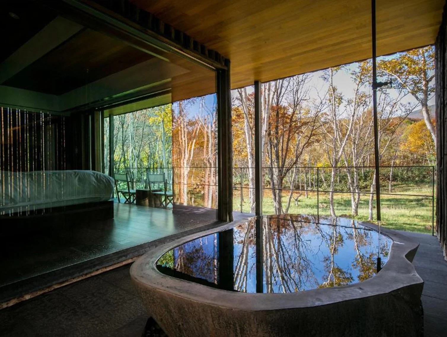 View of a bedroom with a glass wall, overlooking a forest with trees in autumn colors, and a hot tub in the foreground.