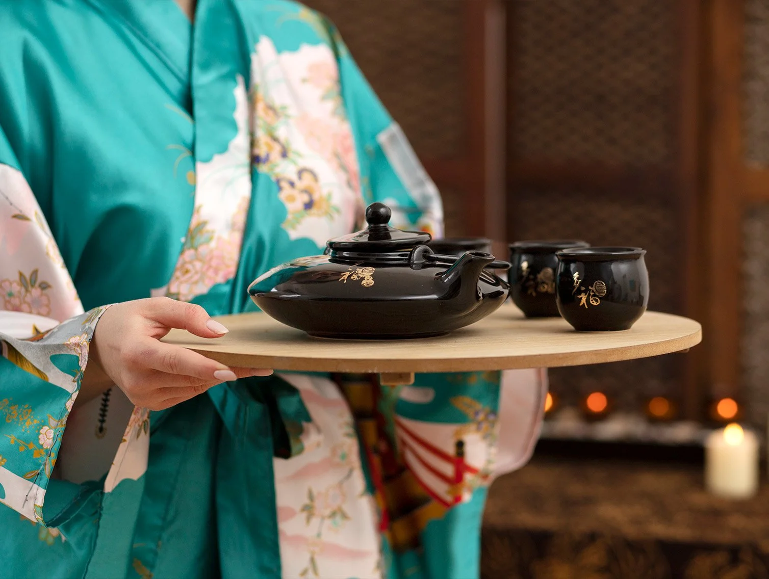 Person in a colorful kimono holding a round wooden tray with a black teapot and three black tea cups.