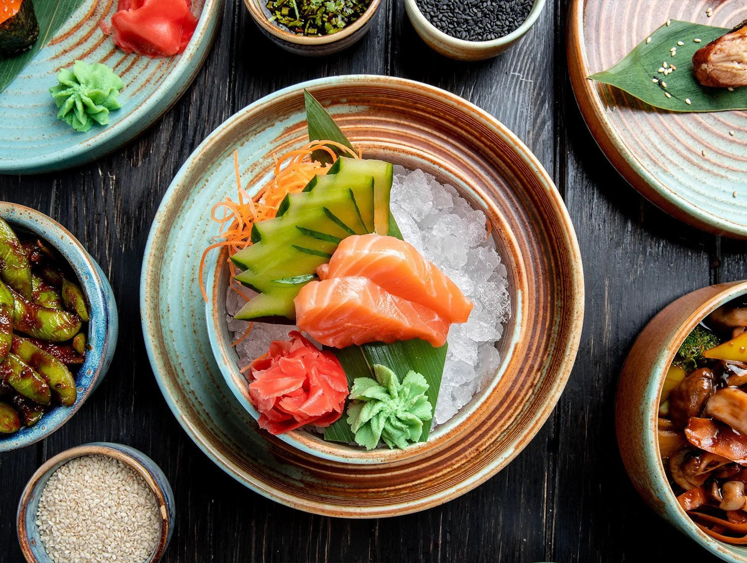 A plate of sushi with slices of salmon, cucumber, shredded carrots, wasabi, and pickled ginger, set on ice. Surrounding bowls contain various side dishes and condiments.