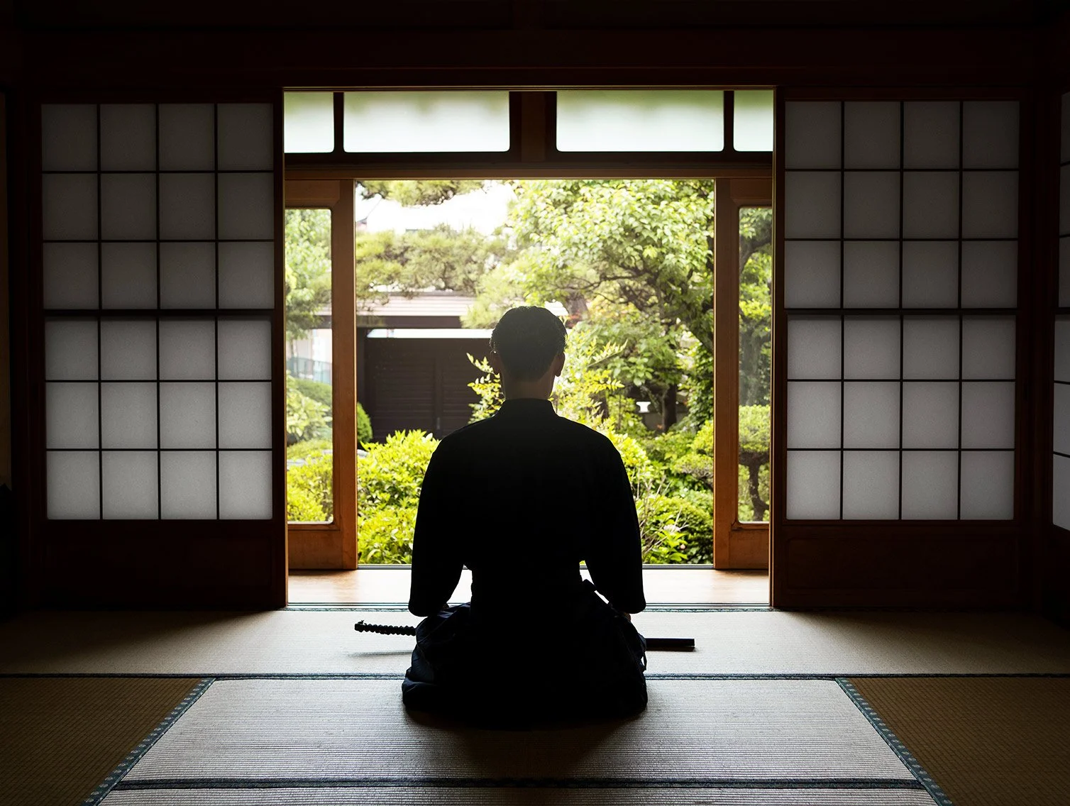 A person dressed in traditional Japanese martial arts attire, sitting in a meditative pose on tatami mats inside a room, looking out at a lush garden through open sliding shoji doors.