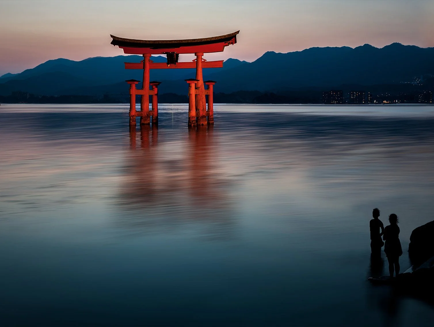 A traditional Japanese torii gate standing in calm water at twilight, with a mountain range in the background and two people standing on the shoreline in silhouette.