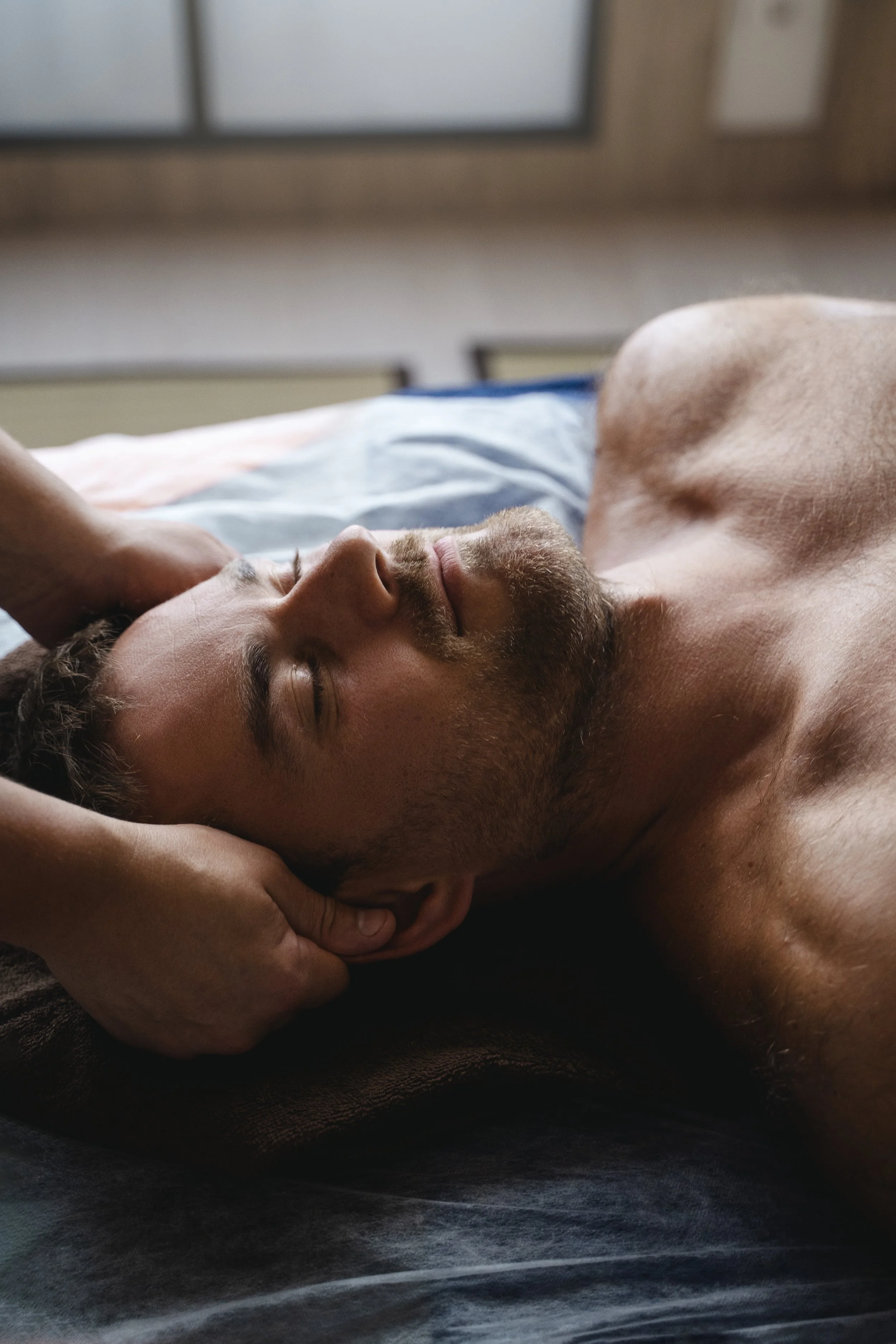 A shirtless man with short hair and a beard lying on his back with eyes closed, resting his head on a pillow, with a hand gently cradling his head.