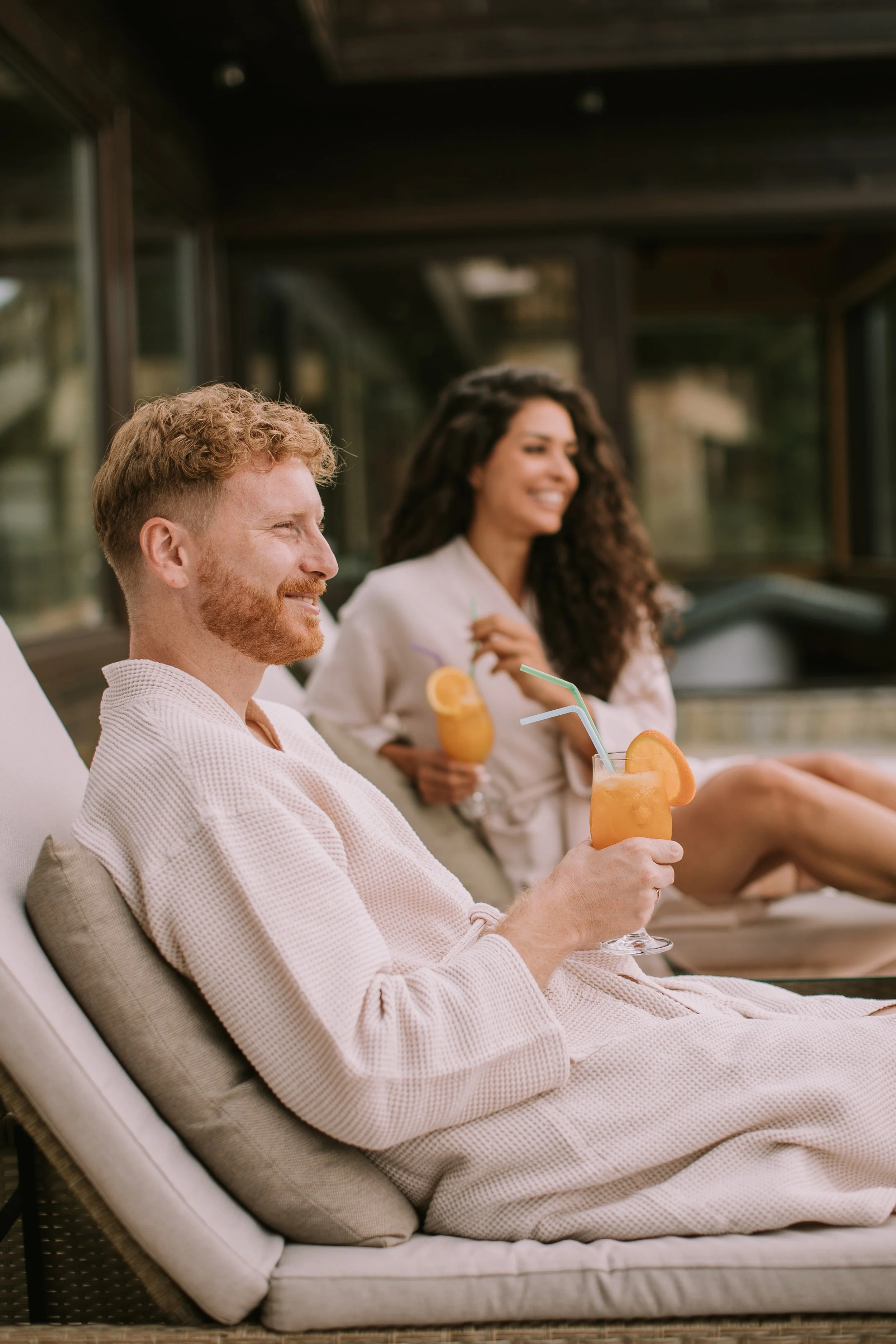 A man and a woman relaxing outside, wearing robes, each holding a drink with a straw and orange slice, smiling and enjoying their time.