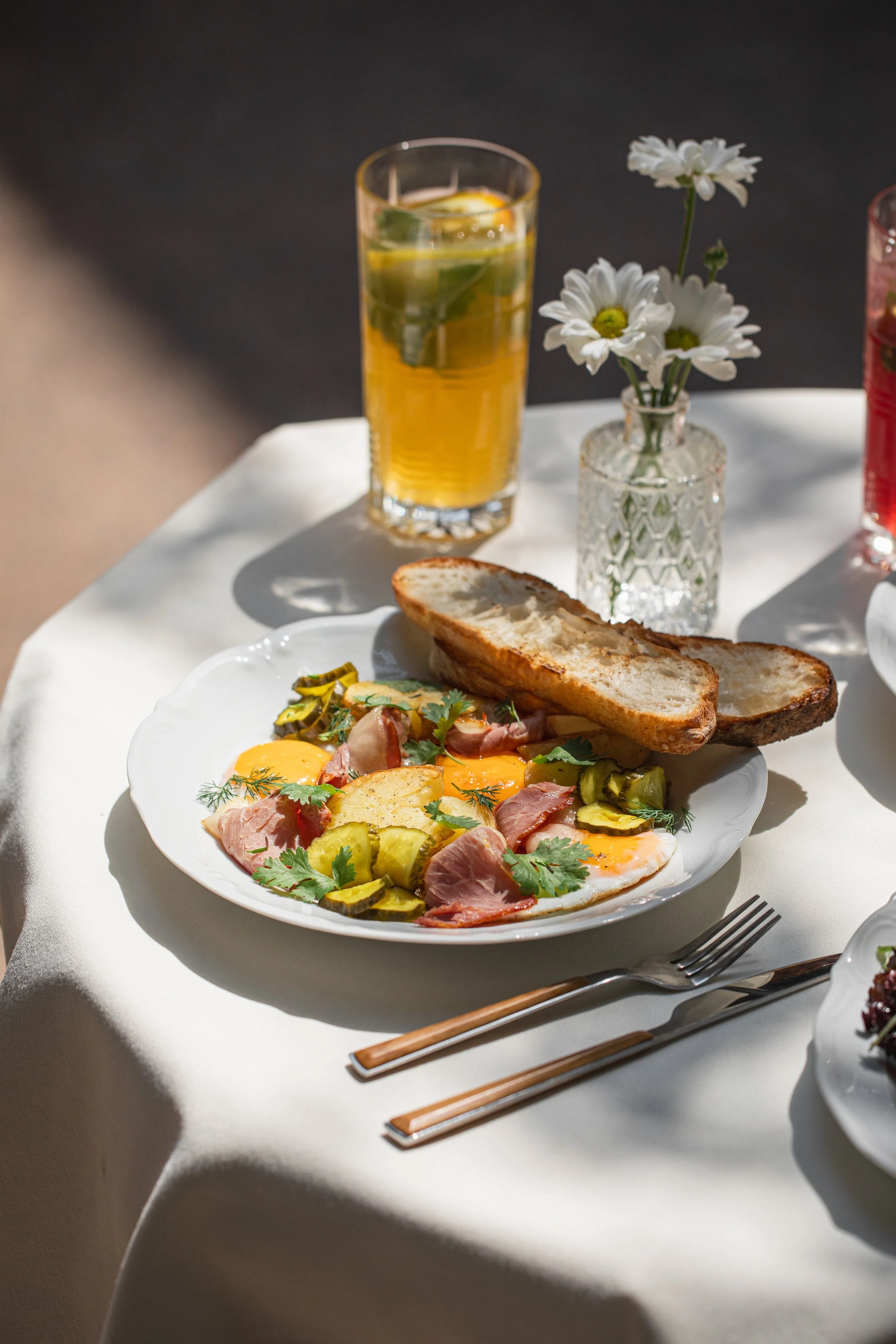 A breakfast plate with fried eggs, ham, pickles, and herbs, accompanied by two slices of toasted bread on a white tablecloth. In the background, a tall glass of iced tea with lemon and mint, a small vase with white daisies, and a fork and chopsticks are visible.
