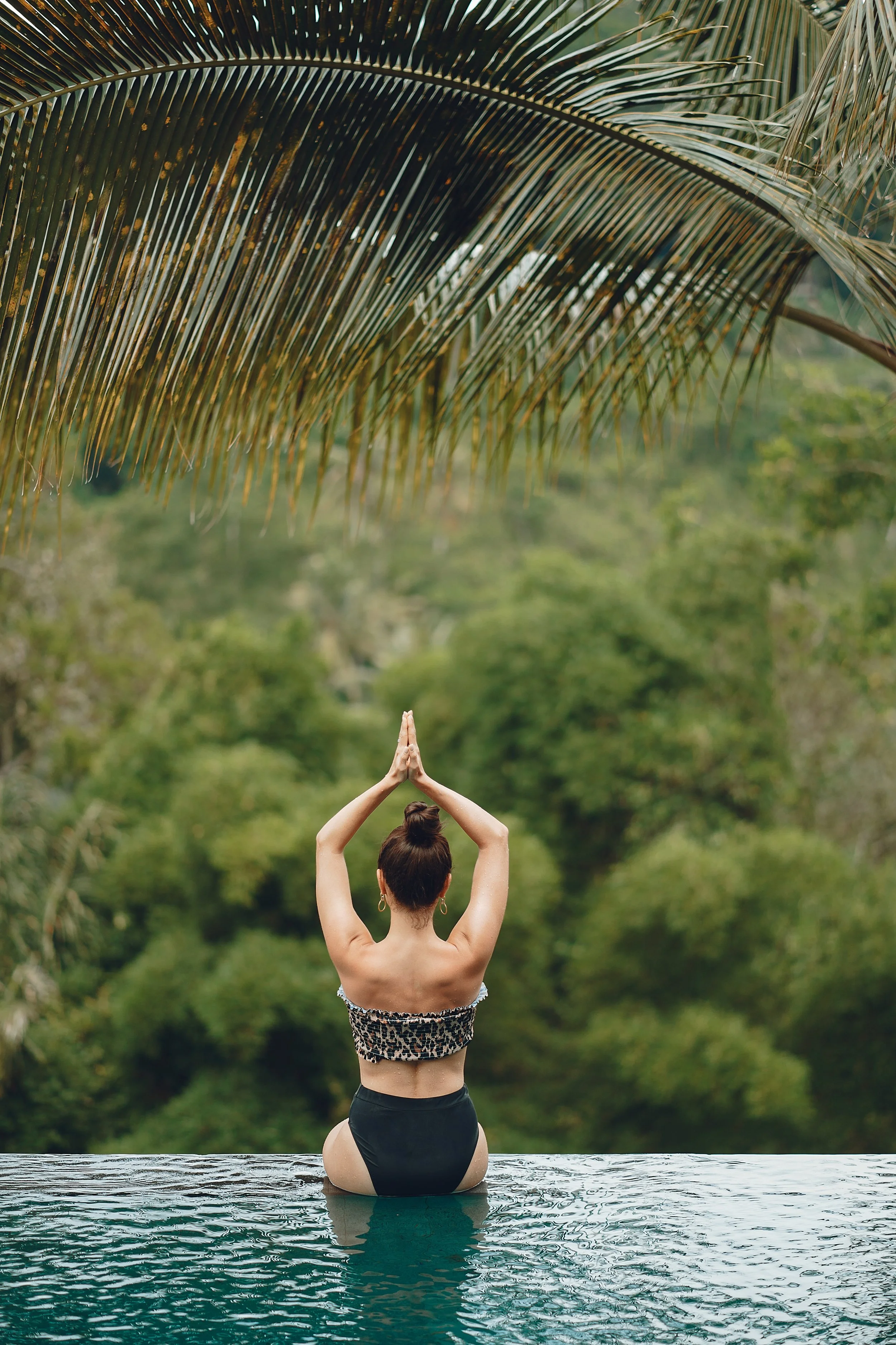 A woman practicing yoga by a pool in a green, tropical setting, facing away with her hands in a prayer position above her head, wearing a strapless top and shorts, with palm leaves overhead.