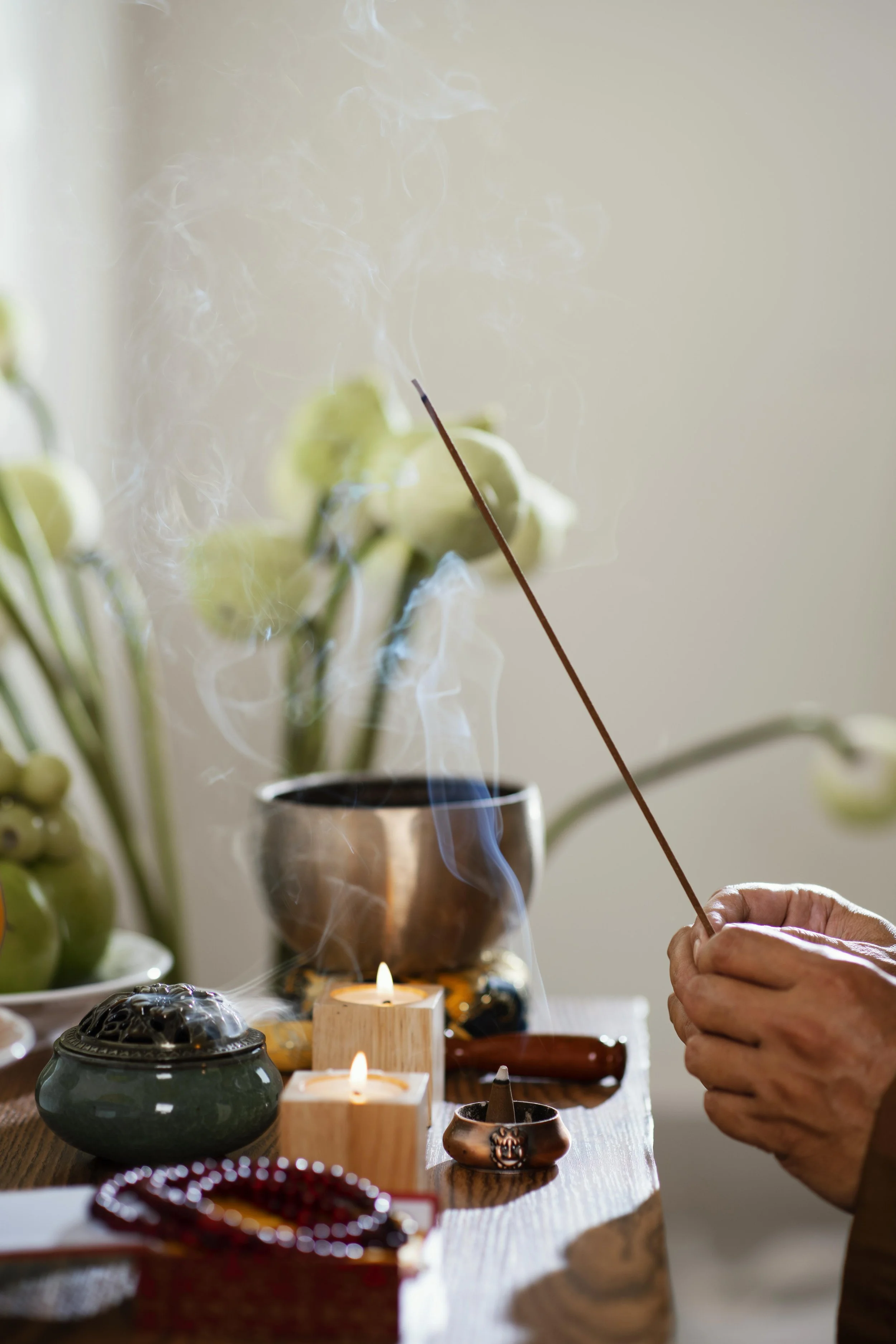 Person holding incense stick near candles and incense burners on a table, with blurred candles and tableware in the foreground.