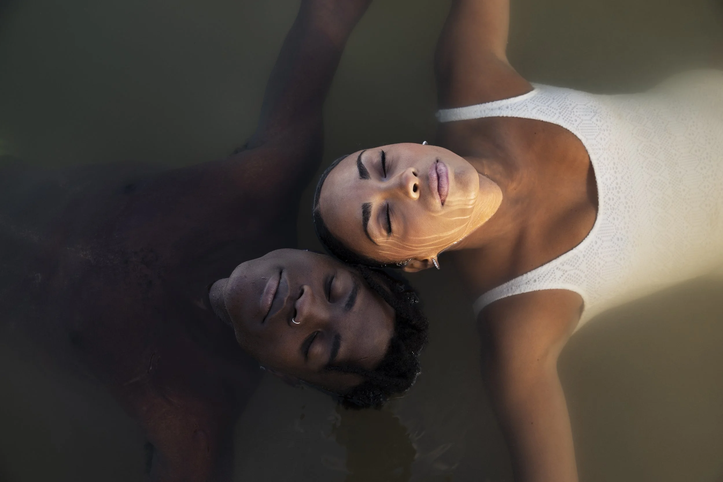 Two women lying with eyes closed, one with darker skin and dark hair, and the other with lighter skin and dark hair, wearing a white dress, close together in water.