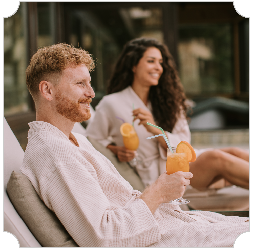A man and a woman relaxing outdoors with drinks, smiling. The man has red hair and a beard, wearing a bathrobe. The woman has curly hair and is holding a similar drink, sitting on lounge chairs.