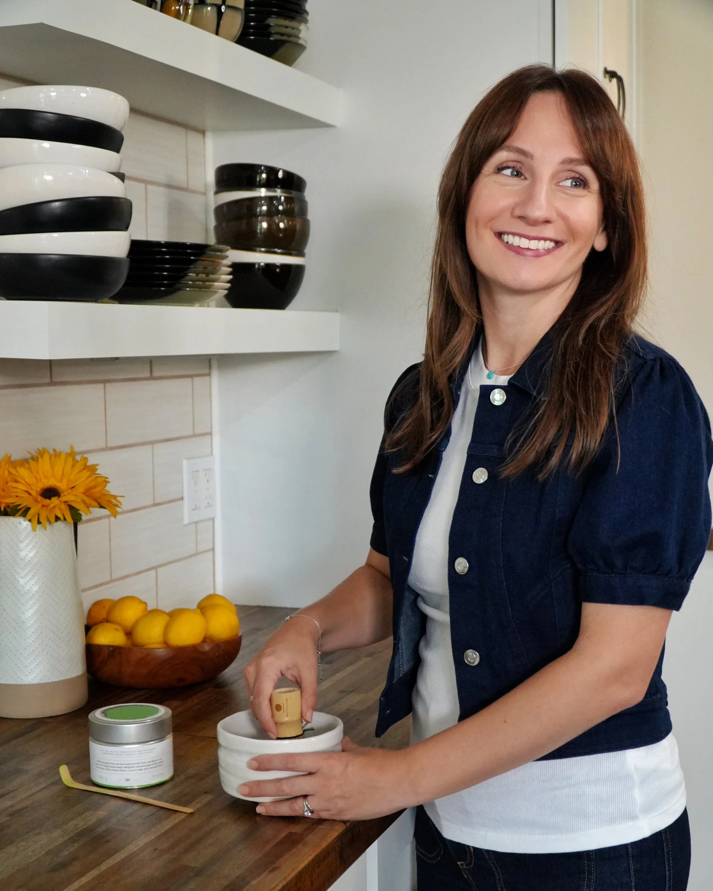 Karman Meyer, RDN in a kitchen, using a wooden pestle to grind ingredients in a white mortar. There are yellow lemons and a small jar on the wooden countertop, with stacked bowls and plates on the shelf behind her.