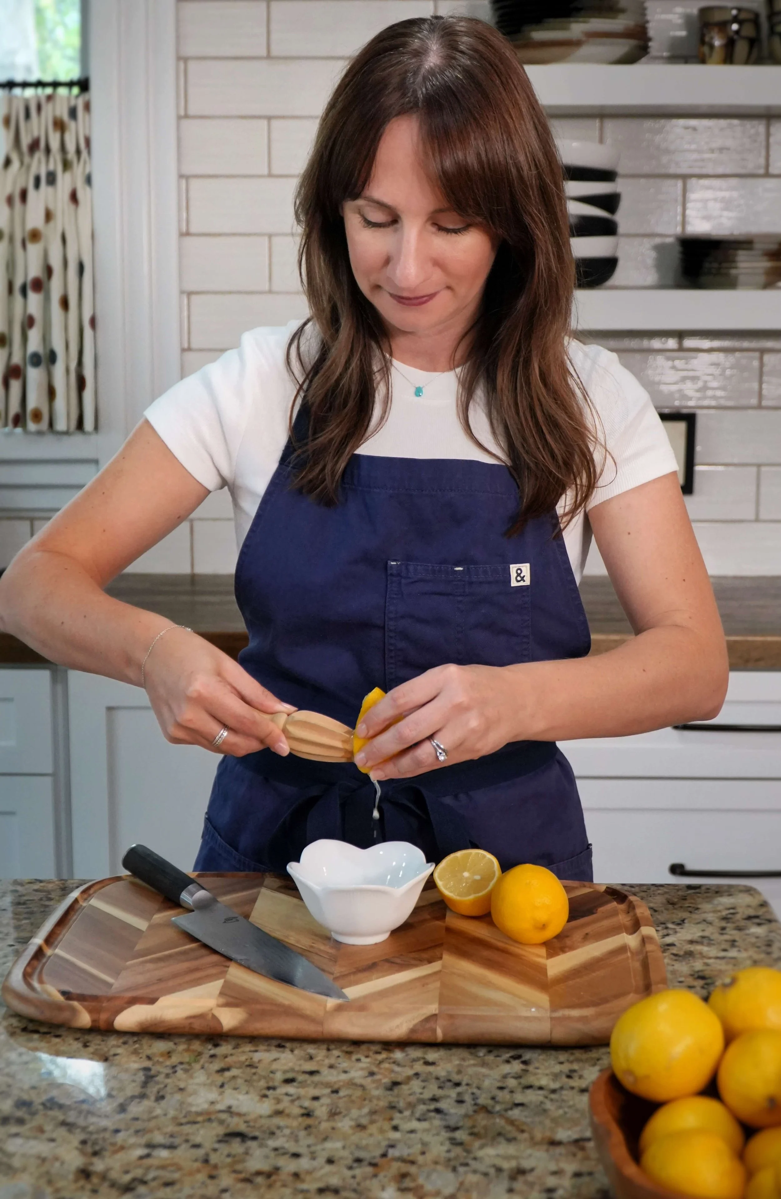 A woman wearing a navy apron and white t-shirt is standing in a kitchen, squeezing a lemon over a cutting board. There are fresh lemons on the cutting board and in a bowl, with a knife and a white dish nearby, in a cozy kitchen setting with white tiled walls.