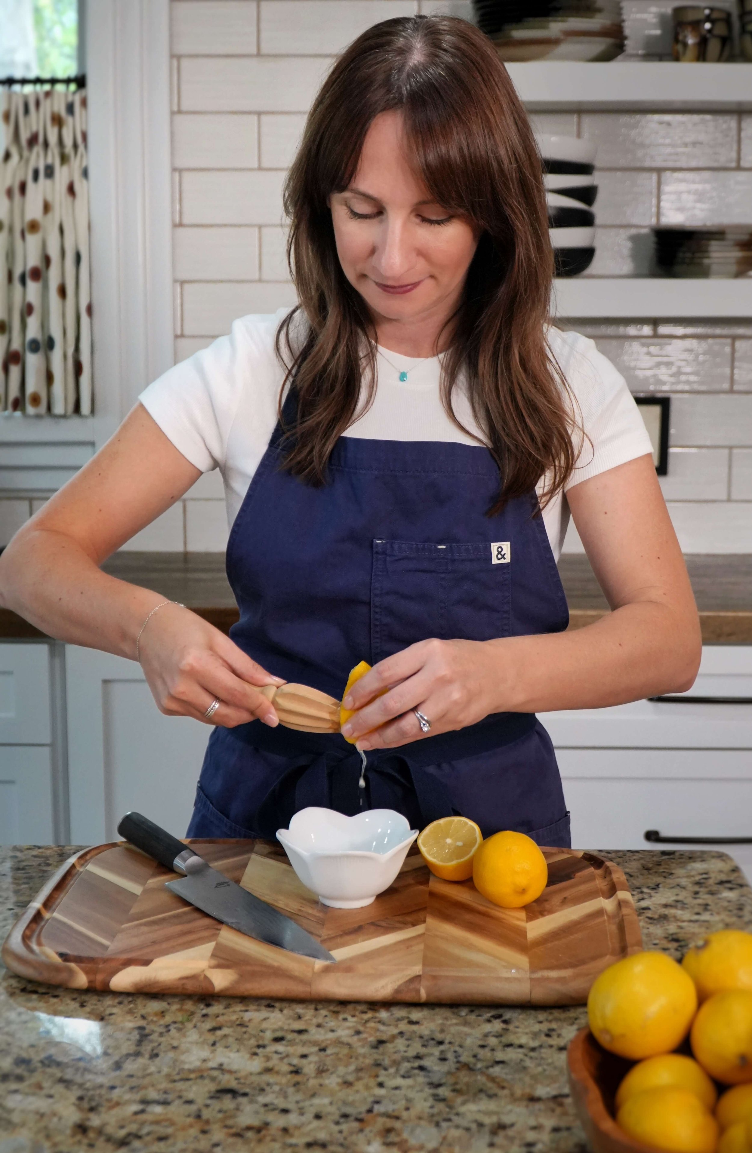 A woman in a white shirt and blue apron is in a kitchen, squeezing a lemon over a white bowl on a wooden cutting board. There are whole lemons and a knife on the counter, and open shelves with dishes and bowls in the background.
