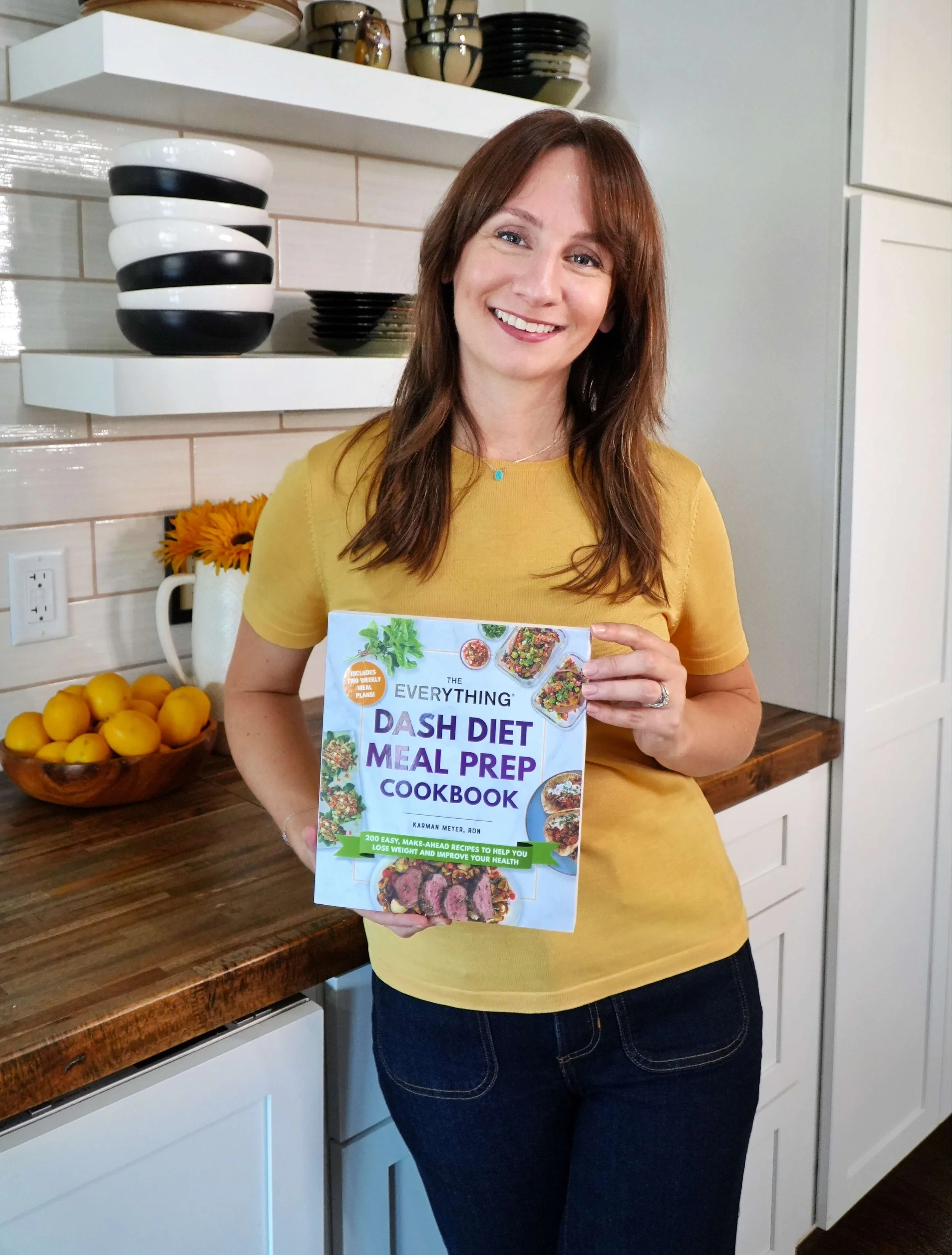 A woman with brown hair smiling and holding a cookbook titled 'Dash Diet Meal Prep Cookbook' in a kitchen. There are bowls and lemons on the counter behind her.