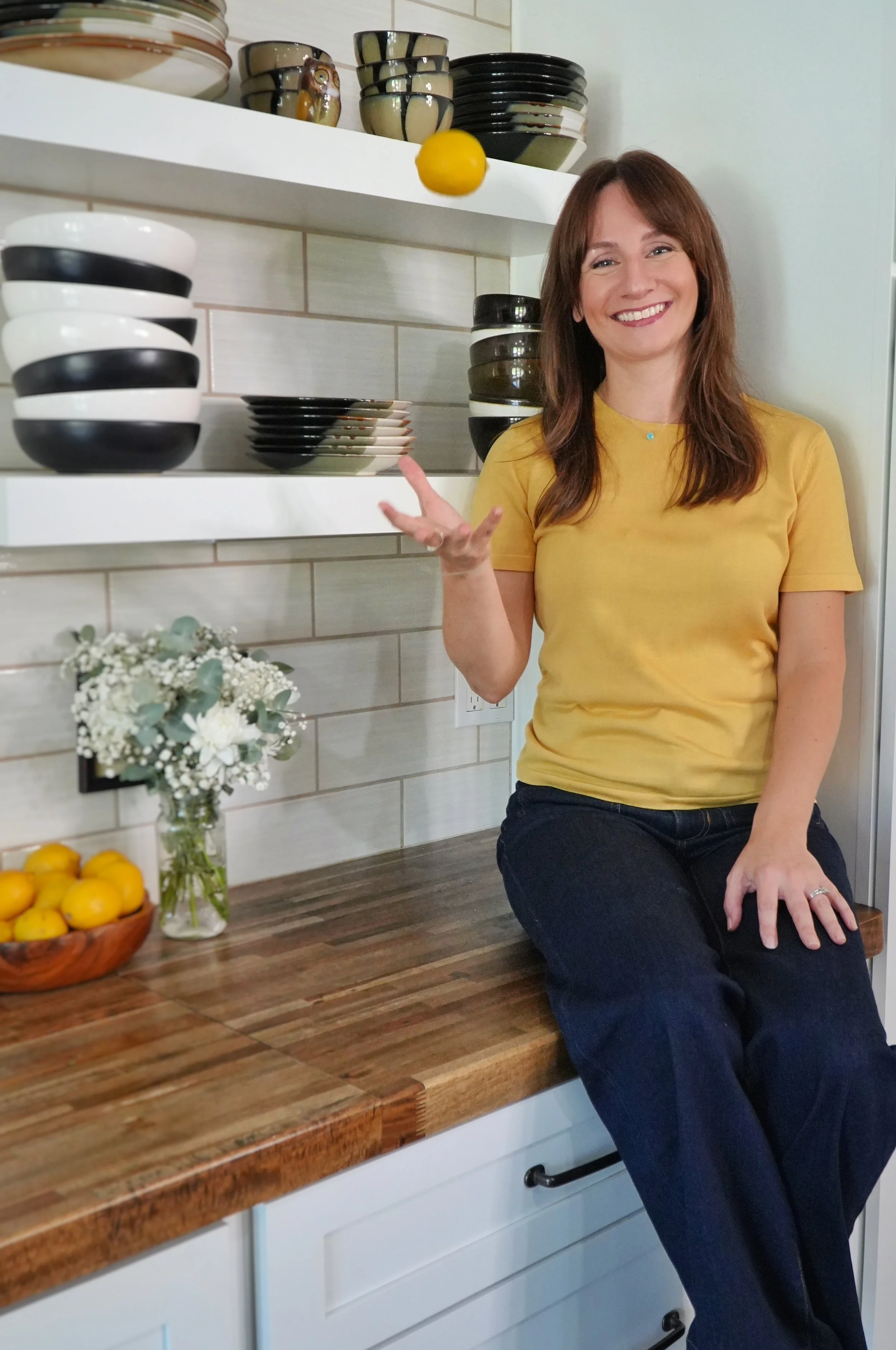A woman with brown hair, wearing a yellow shirt and dark pants, sitting on a kitchen counter next to a vase of white flowers, a bowl of lemons, and a shelf with stacked bowls and plates.