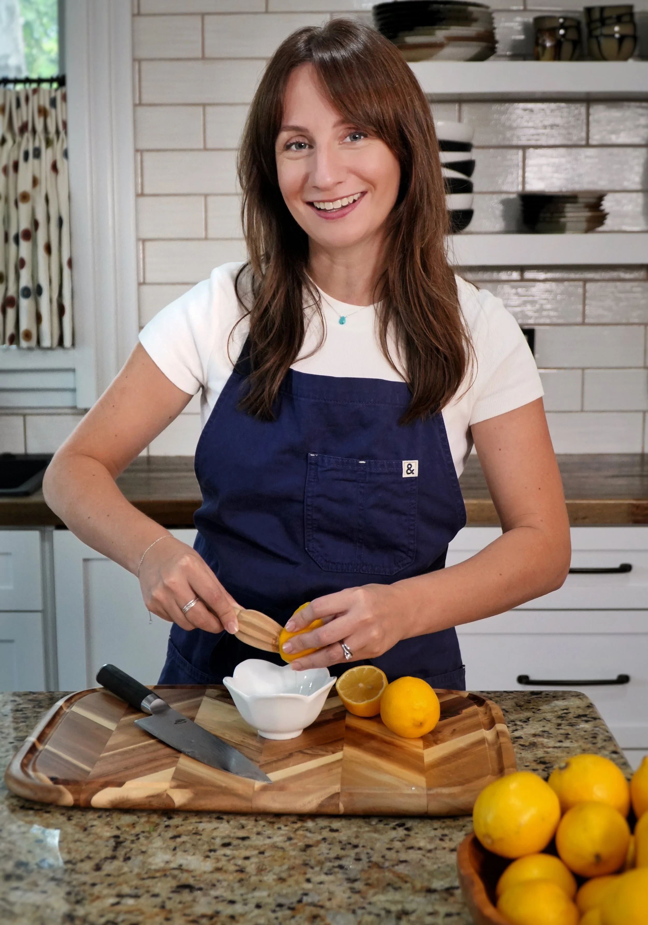A woman with long brown hair wearing a white t-shirt and a navy apron is preparing lemons in a kitchen. She is holding a lemon and using a zester. The kitchen has a stone countertop, a wooden cutting board, yellow lemons, and a white bowl. There are cabinets and a shelf with dishes in the background.