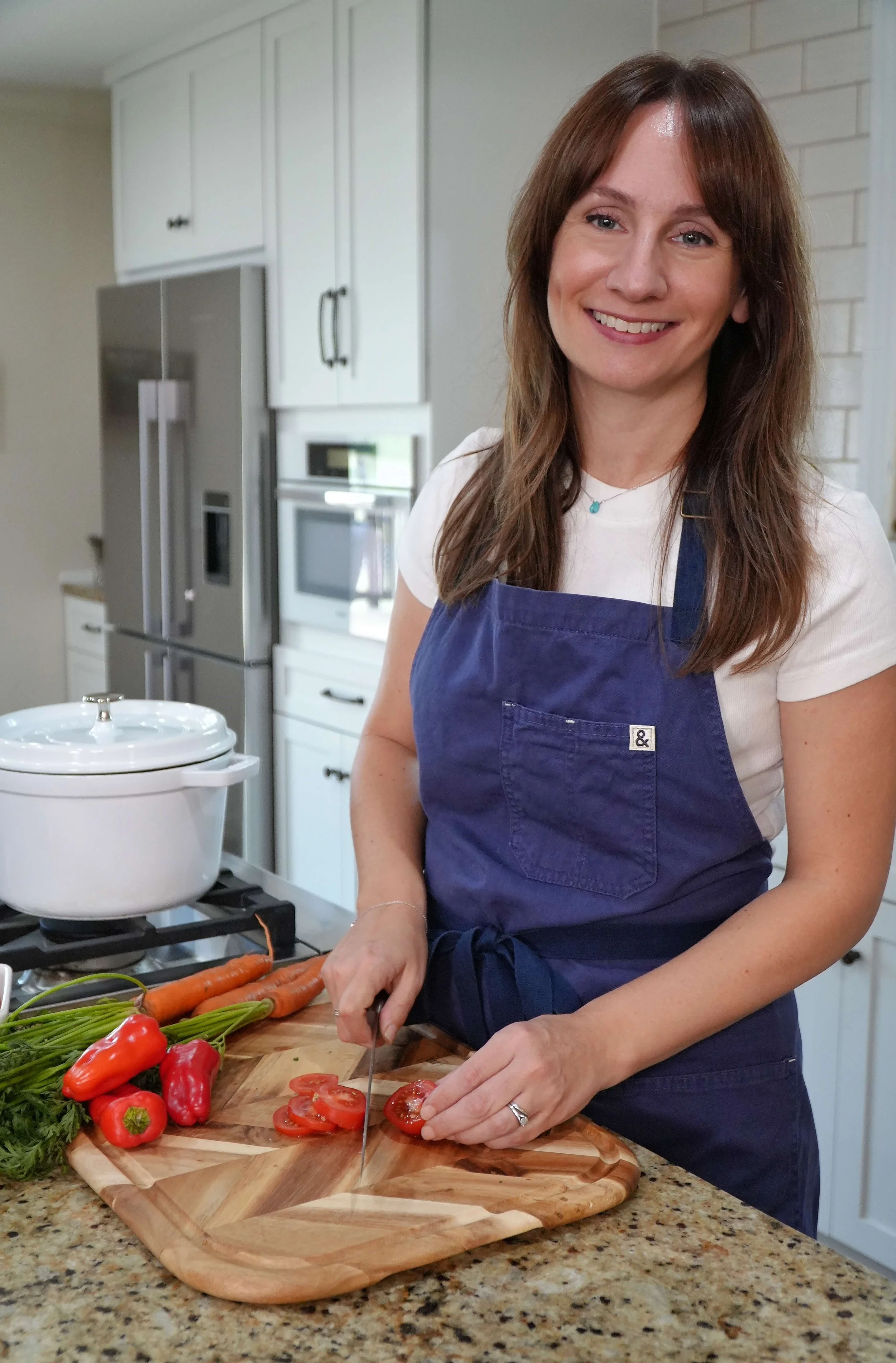 A woman with brown hair and a light complexion is smiling while slicing tomatoes on a wooden cutting board in a kitchen. She is wearing a navy blue apron over a white shirt, and has a silver ring on her left ring finger. The kitchen has white cabinets and a stainless steel refrigerator in the background. There are fresh vegetables like red bell peppers and carrots on the counter.