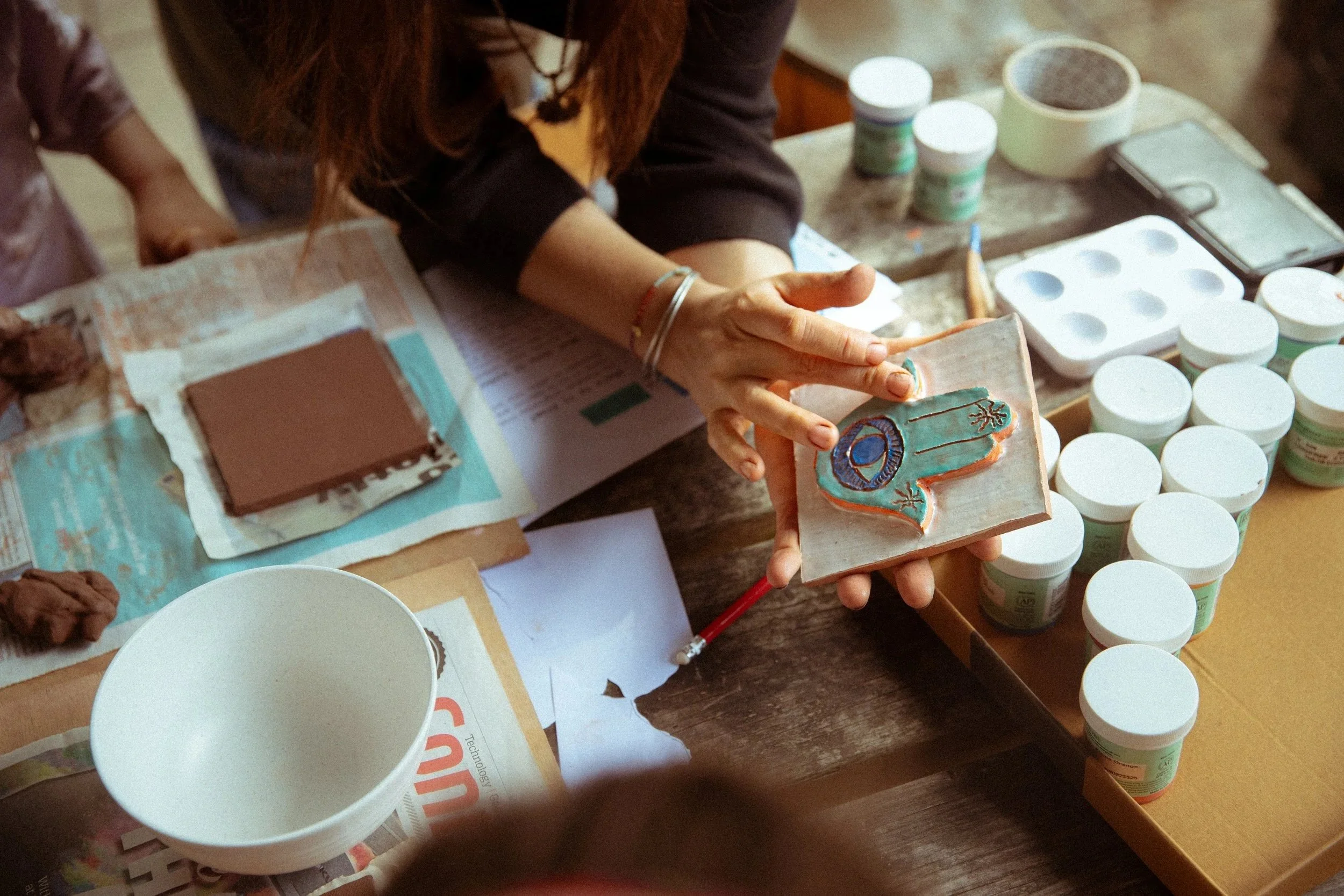 A person holding a decorated ceramic tile with blue, orange, and black designs, surrounded by containers of paint or glaze, on a cluttered table with paper and cups.