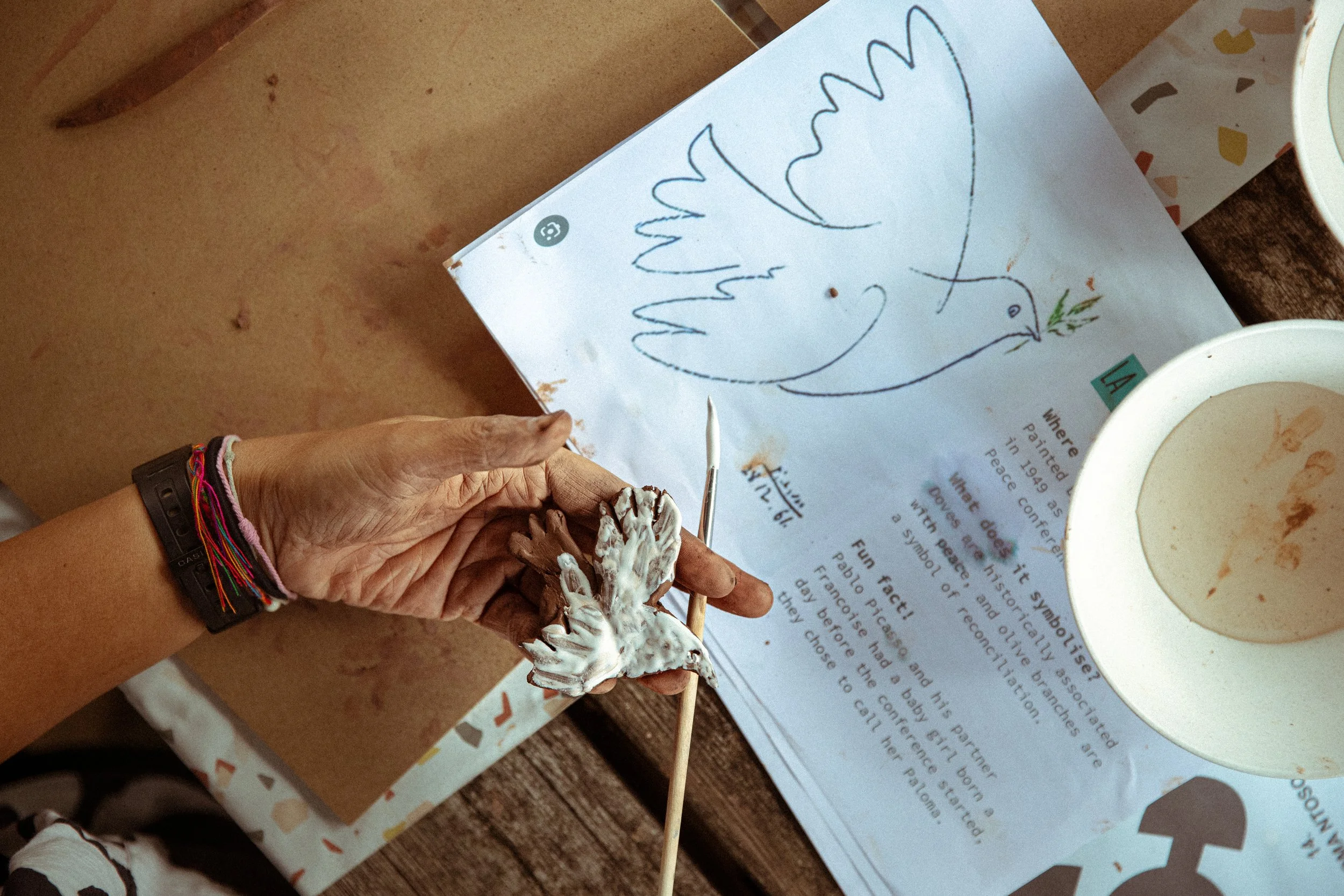 Close-up of a person's hand holding a small paper sculpture of a dove with an olive branch, near a printed page and a cup of coffee on a wooden table.