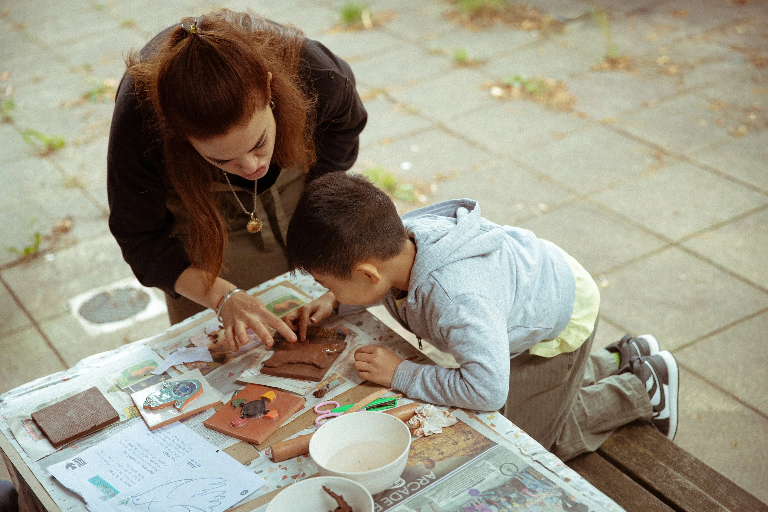 A woman and a young boy are engaged in a craft activity on a table covered with newspapers. The woman has long brown hair and is looking down at the table, while the boy, wearing a gray hoodie, is kneeling and focused on the crafts. Various craft supplies like scissors, paper, and small decorative items are scattered on the table. The scene appears to be outdoors on a paved surface with some weeds growing between the tiles.