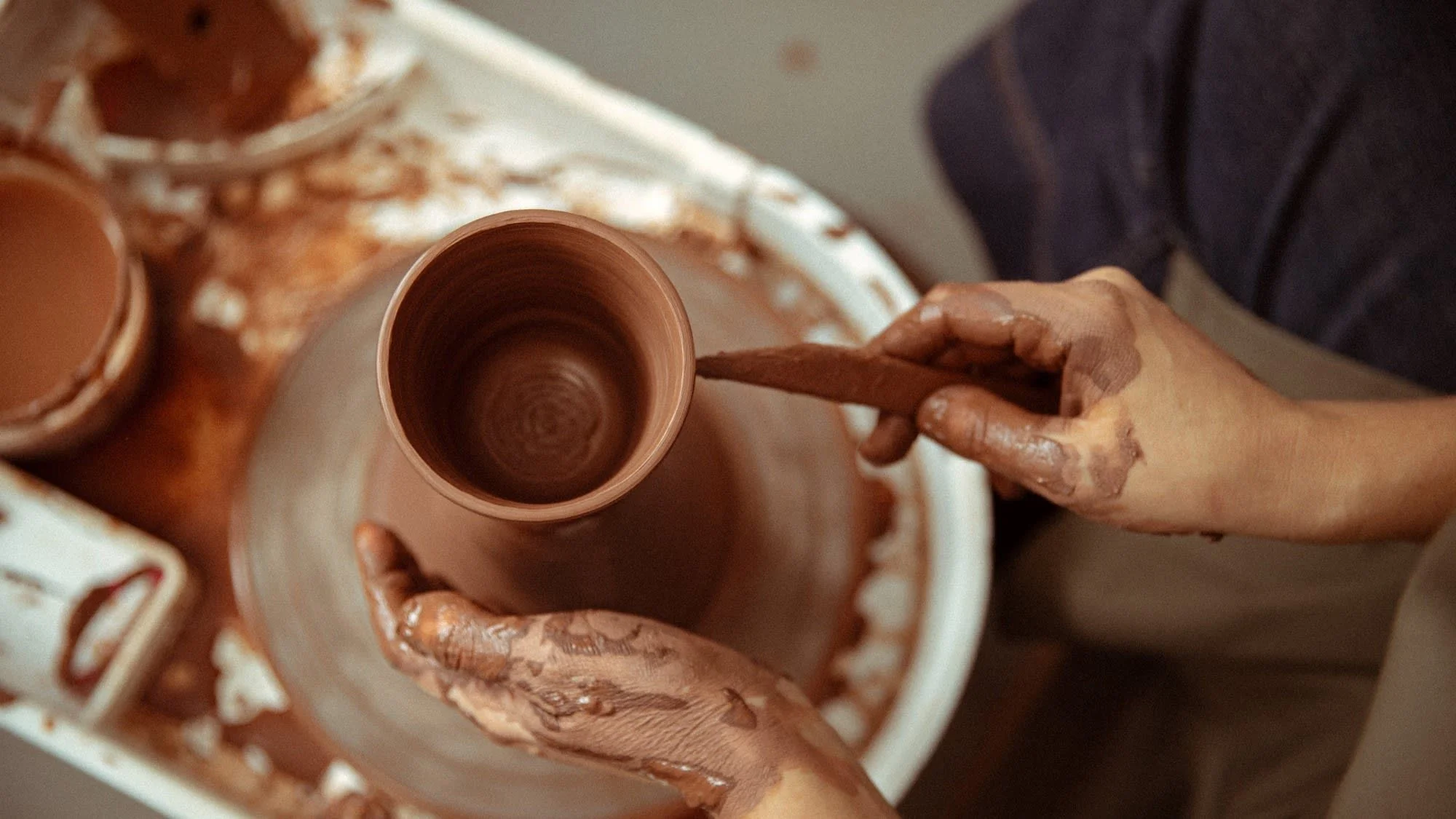 A person shaping a clay pottery piece on a pottery wheel, with hands covered in wet clay.