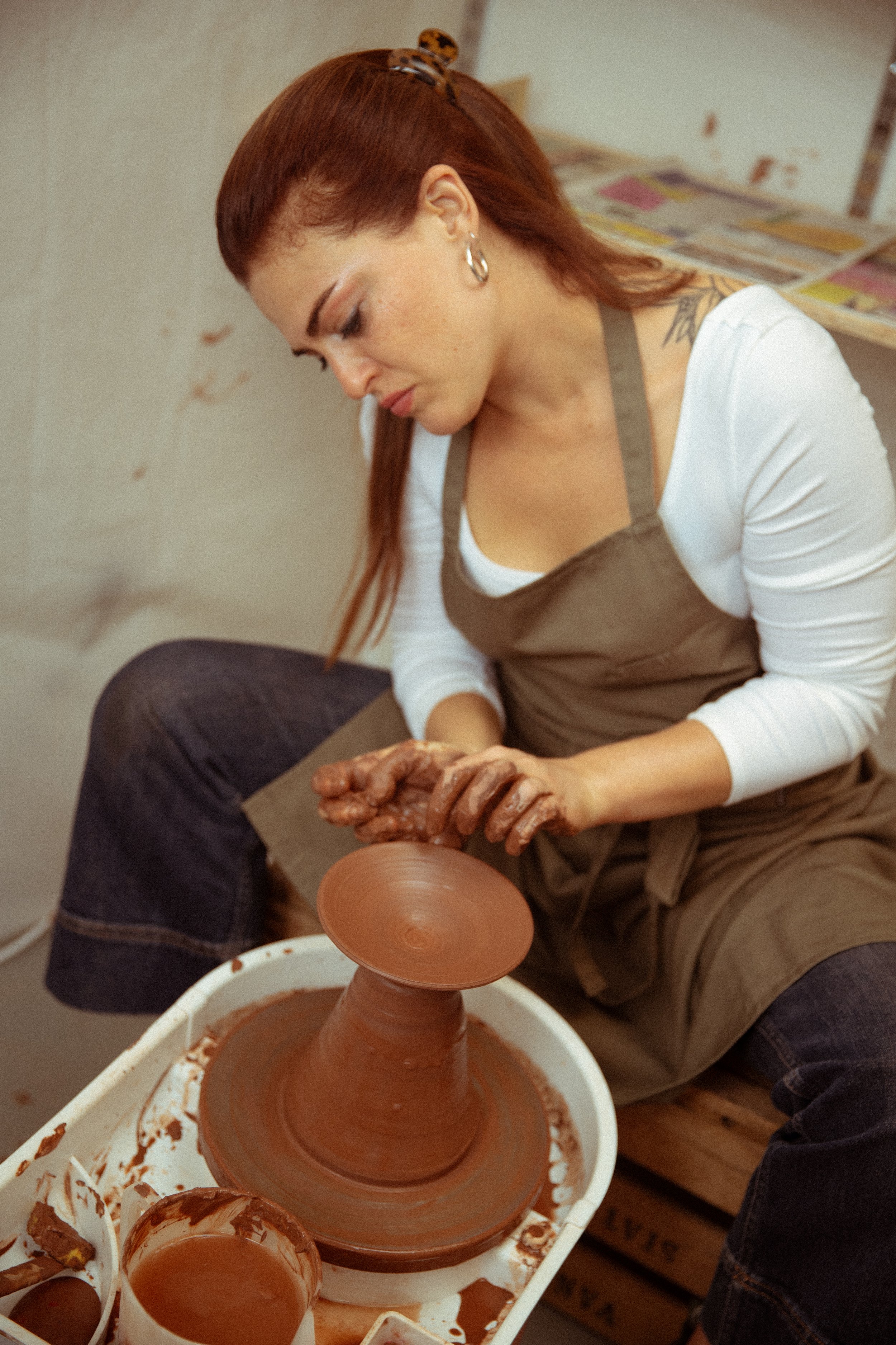 A woman with red hair, earrings, and a tattoo on her shoulder is working on a pottery wheel to shape a clay vase. She is wearing a white shirt and a brown apron, and the workspace has magazines or papers on the background wall.