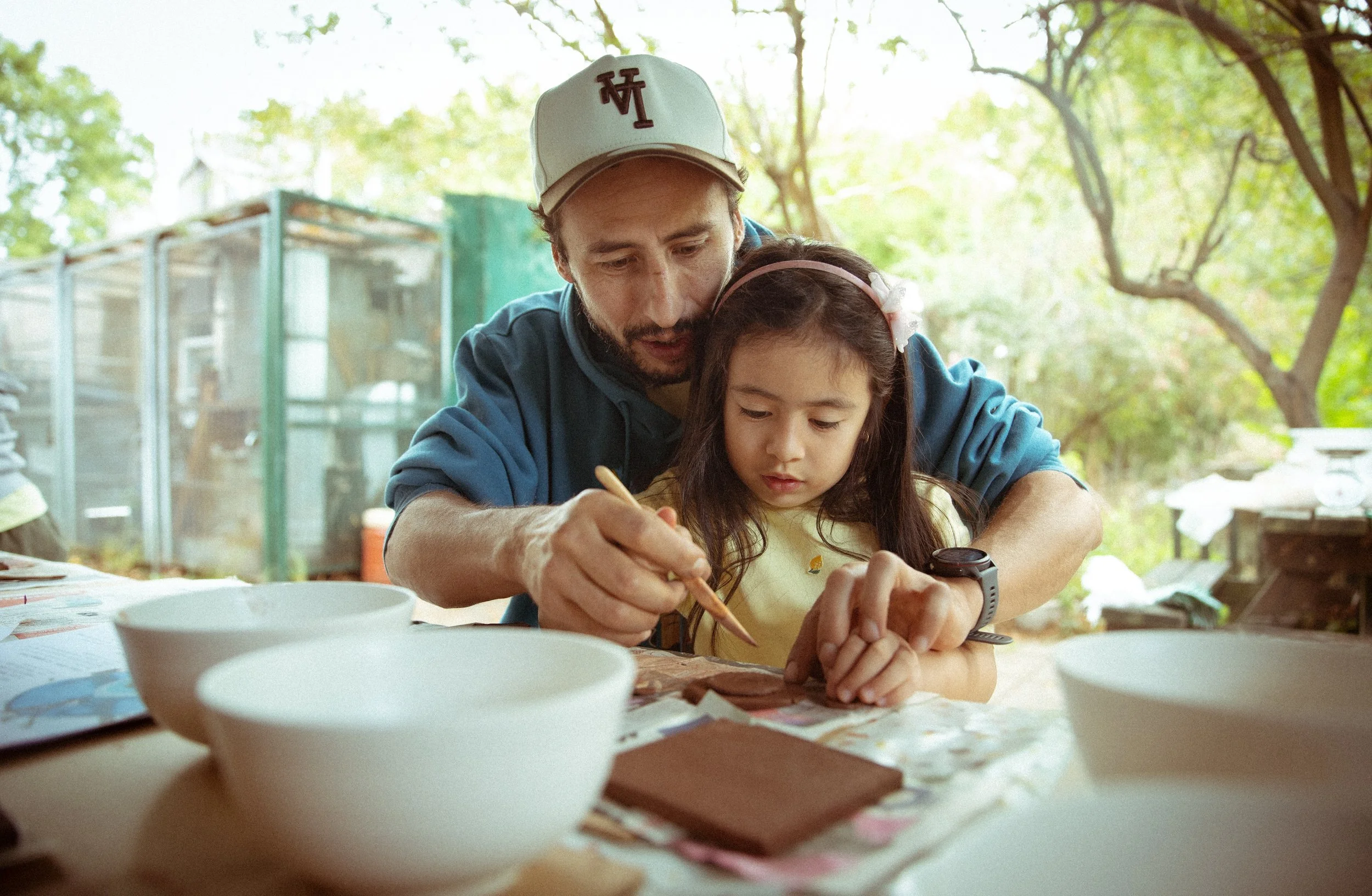 A man and young girl sitting at a table outdoors, engaged in an activity, surrounded by trees with dappled sunlight, with bowls and papers on the table.