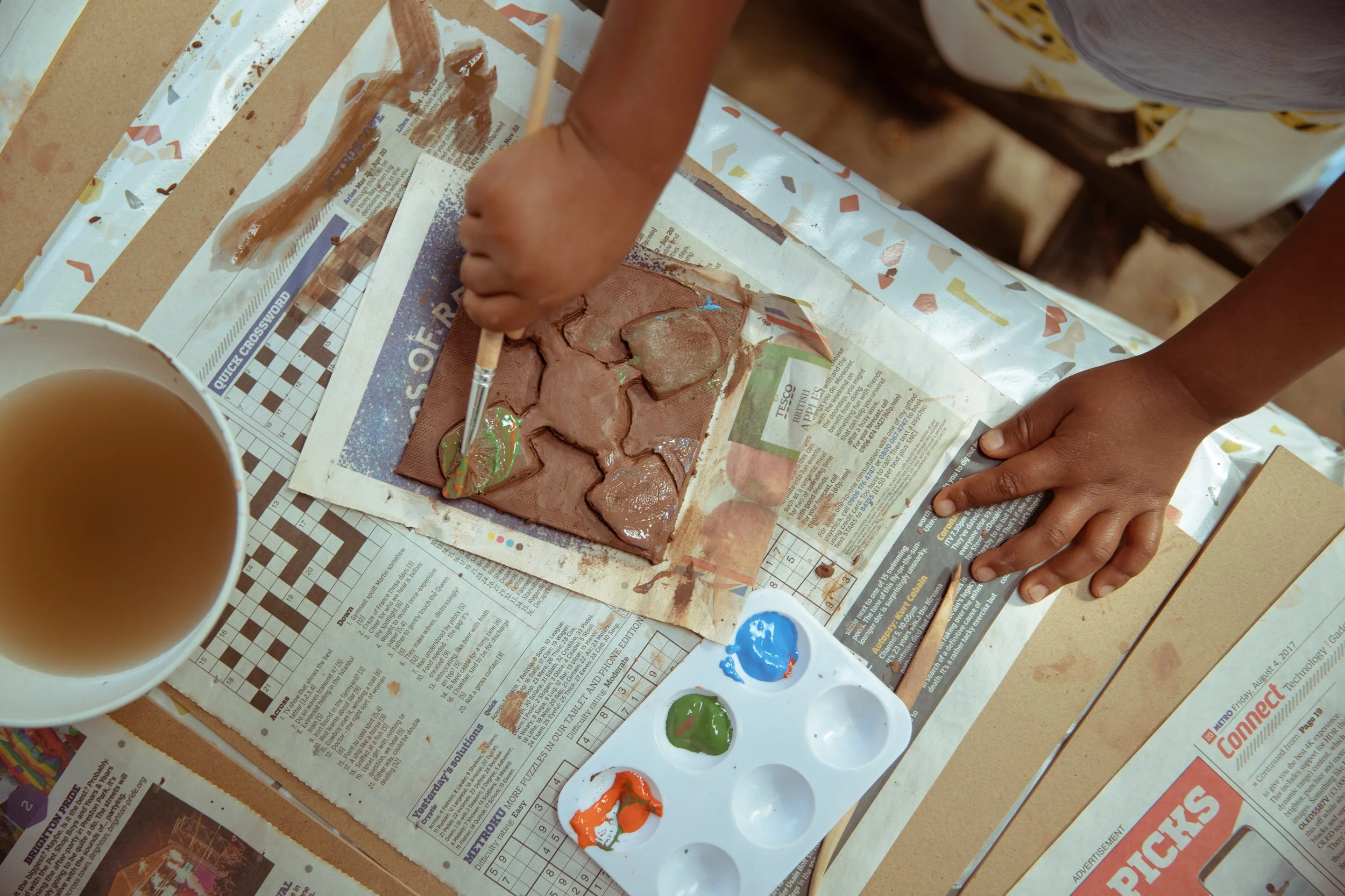 Child painting on a brown textured surface with green, orange, and blue paint using a small brush. The surface is on top of a newspaper-covered table with art supplies scattered around, including a cup of tea or coffee and a paint palette with various colors.
