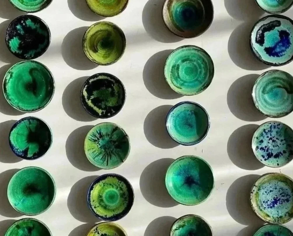 Collection of small round glass jars with various green, blue, and black abstract and swirling designs, arranged on a white surface with shadows cast around them.