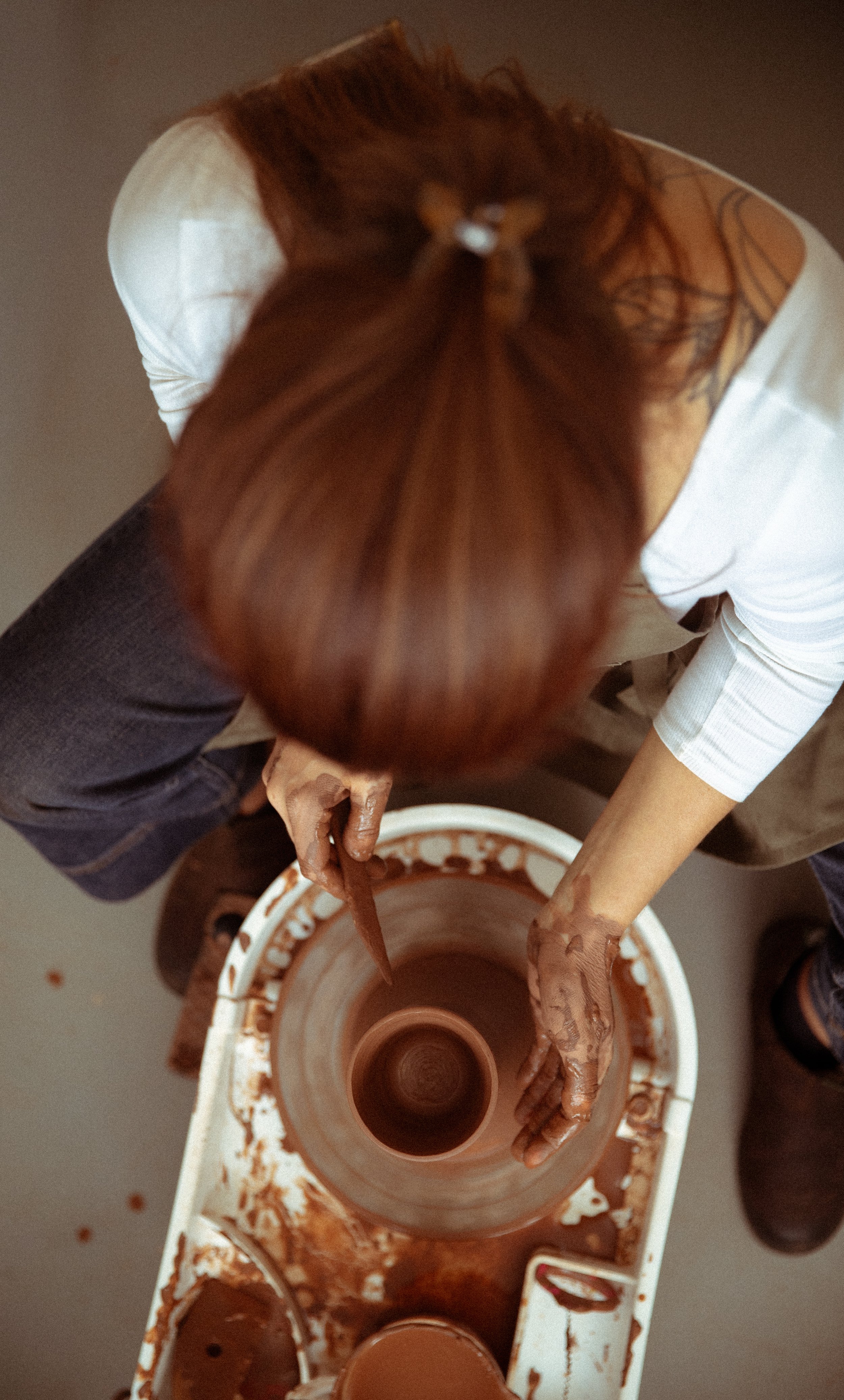 A person with brown hair in a ponytail and tattoos on their shoulder is using a pottery wheel to shape clay. The person has their hands in the clay and is working with a small amount of water near the wheel. The pottery wheel is covered in wet clay and appears to be in a studio.