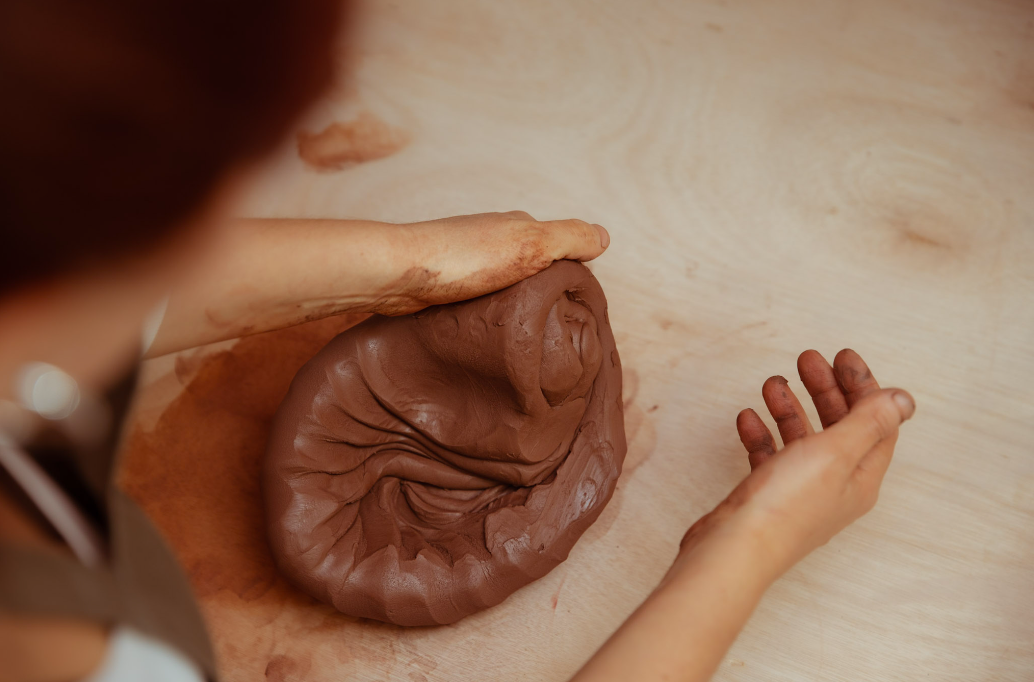 Close-up of a hand shaping a sculpture made of brown clay on a wooden surface.