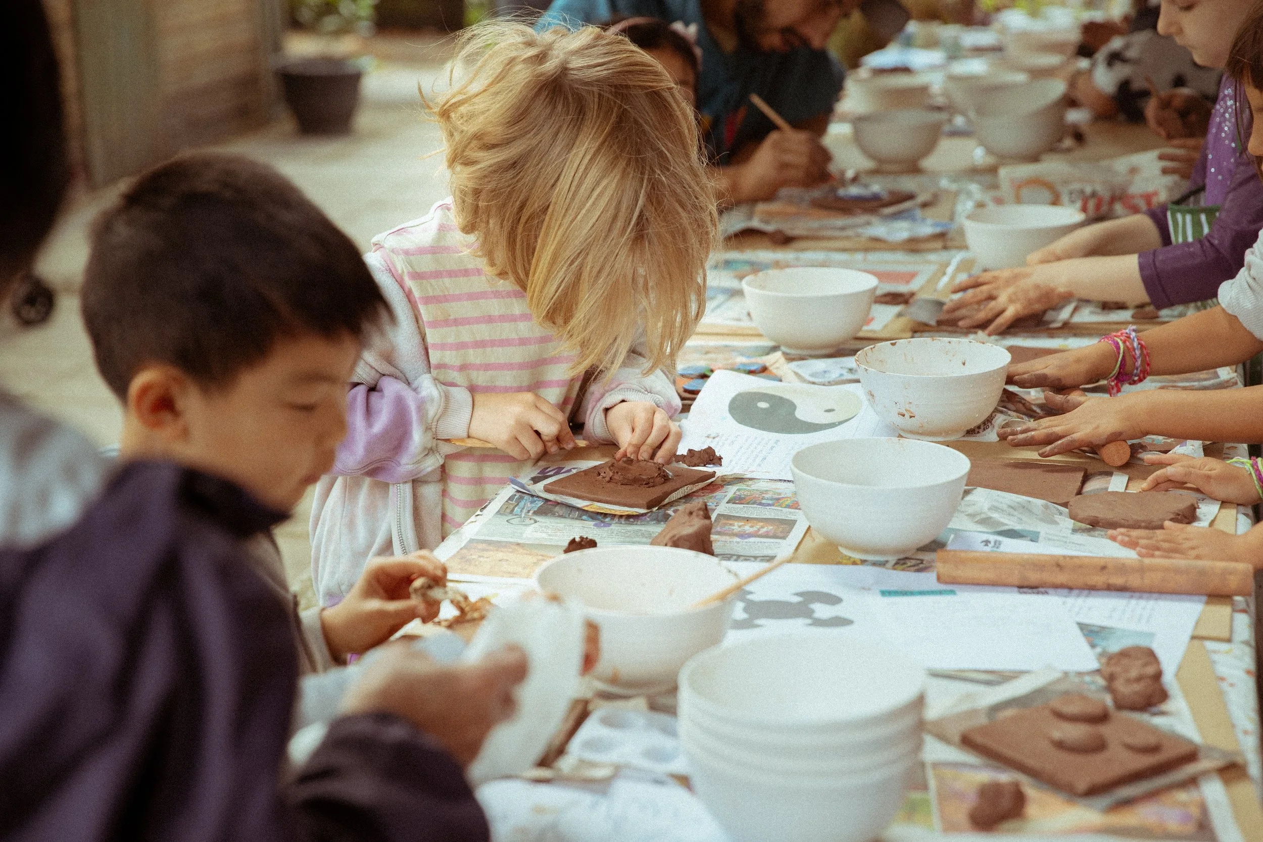 Children participating in a clay sculpting activity at a table covered with newspapers, with bowls and clay materials.