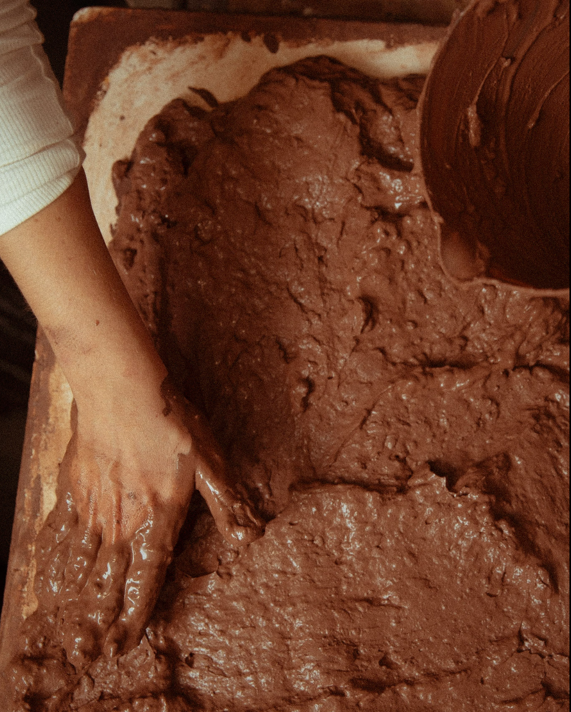 A person mixing thick chocolate dough on a wooden surface with a rolling pin and their hand covered in dough.