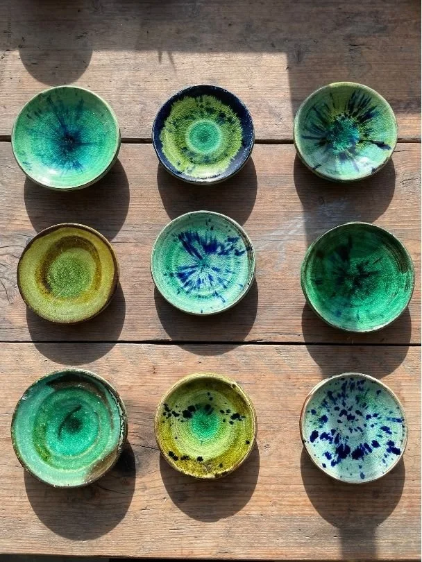 Nine small ceramic bowls with green and blue glaze patterns, arranged in a 3x3 grid on a wooden surface with sunlight casting shadows.