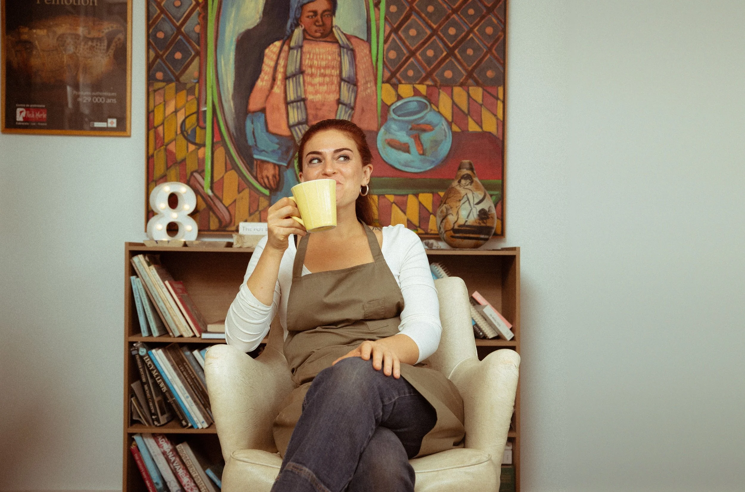 A woman with reddish-brown hair sitting in a cream armchair, holding a yellow mug, wearing a white long sleeve shirt and a brown apron. Behind her, there is a bookshelf filled with books, a large colorful painting of an Indigenous woman holding a basket, two decorative pottery vases, and a white number 8 lamp. The setting appears to be a cozy indoor space.